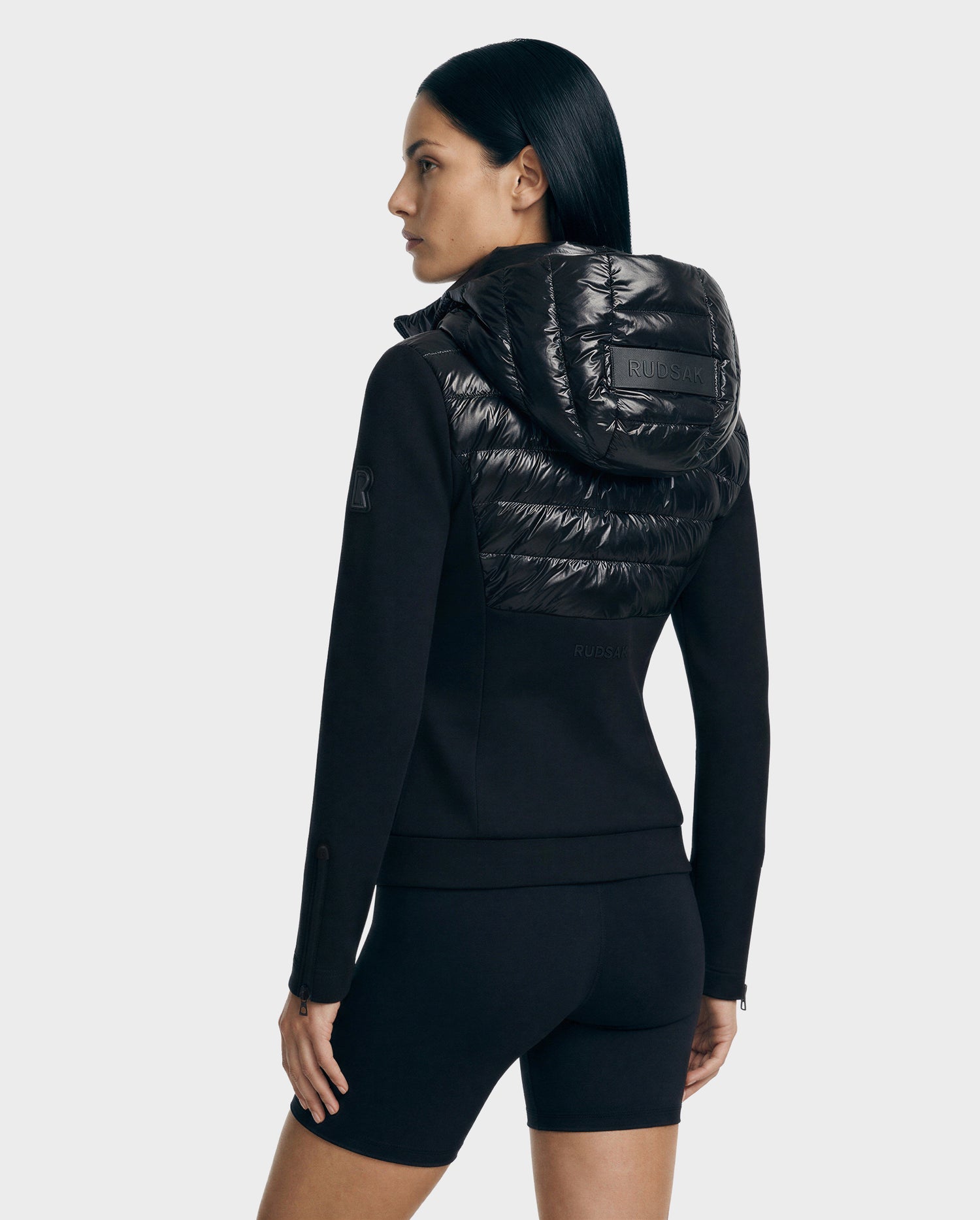 A woman with straight dark hair models the Rudsak MABS C - 8125701 Black fitted jacket with a shiny quilted hood and matching black shorts, standing slightly turned to the side against a light gray background.