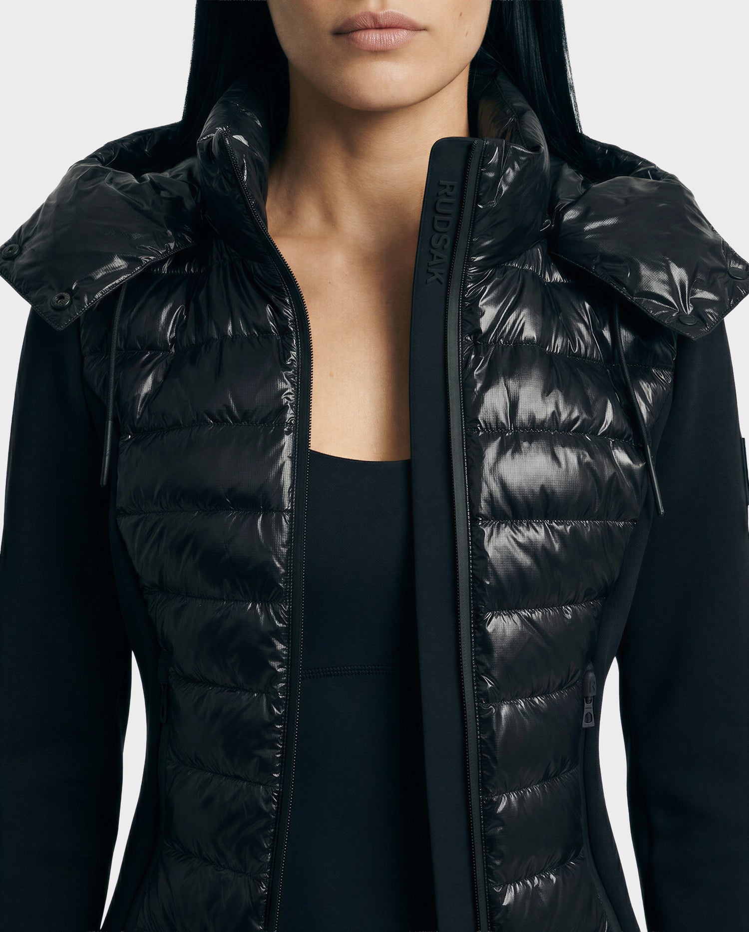 A woman wears the Rudsak MABS C - 8125701 Black puffer jacket, featuring shiny quilted panels and a high collar over a black top, shown from the chest up against a plain background.