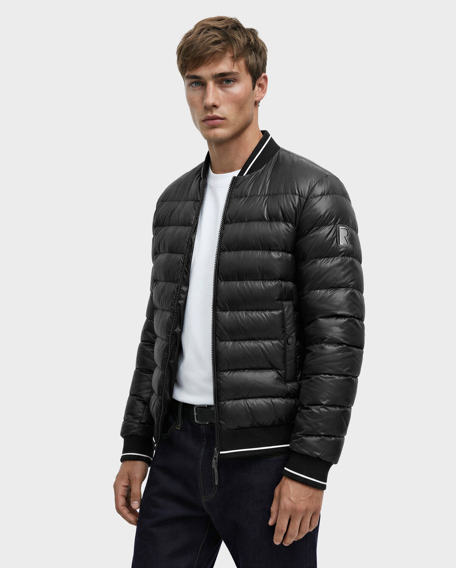 A young man wears the Rudsak JEREMY - 6125025 Black men’s down bomber jacket with black quilted insulation and white trim, paired with a white t-shirt and dark jeans, standing against a light gray background.