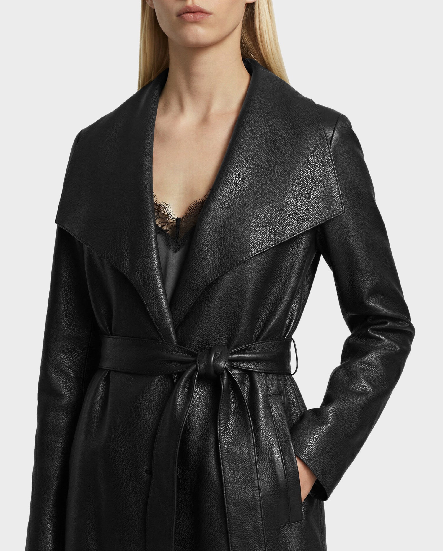 A woman wears the Rudsak MURMU X - 8125670 Black leather trench coat with a wide collar, cinched waist, and lace-trimmed black top—her right hand rests casually in her pocket for a polished, chic look.