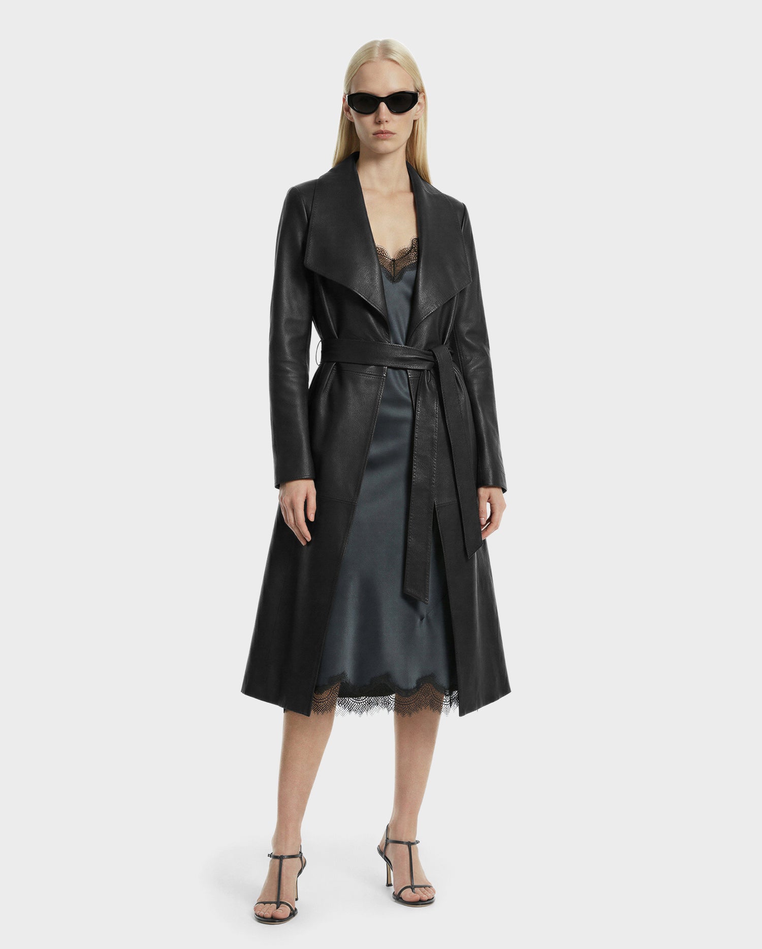 A woman models investment dressing in the Rudsak MURMU X - 8125670 Black long leather coat, styled with black sunglasses, a silky lace-trim slip dress, and strappy heels against a plain light background.