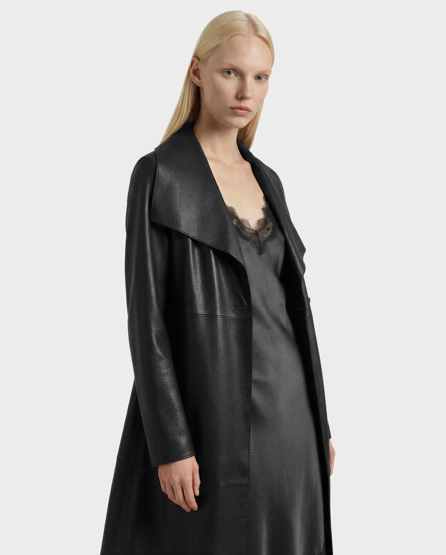 A woman with straight blonde hair models the Rudsak MURMU X - 8125670 Black leather coat over a black lace-trim slip dress, standing against a plain light gray background.