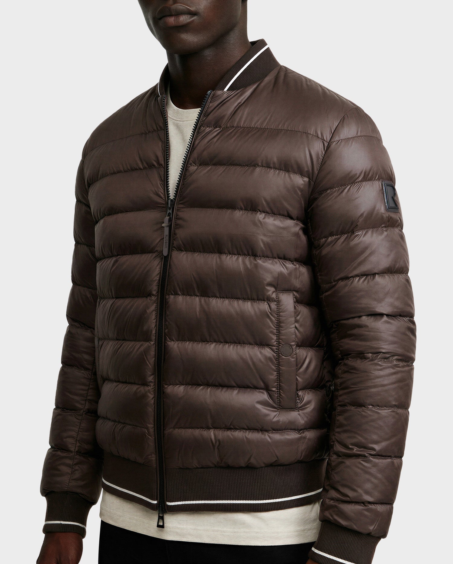 A person models the Rudsak JEREMY - 6125025 Espresso men’s down bomber jacket, featuring quilted insulation, front zipper, ribbed cuffs and hem, and a sleeve patch, worn over a light shirt against a plain background.