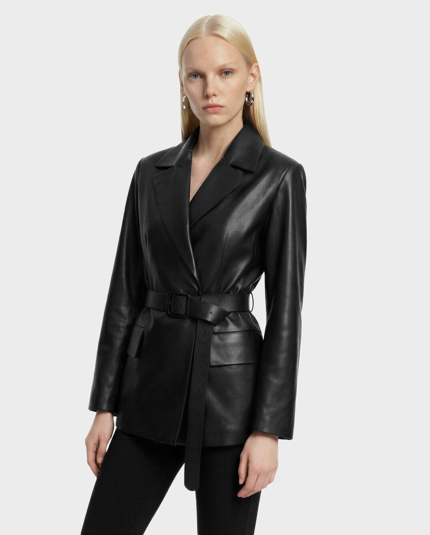 A woman with long blonde hair models the Rudsak KENDALL - 8125617 Black women's leather blazer with a sleek black belt over black pants, standing against a plain light gray background.