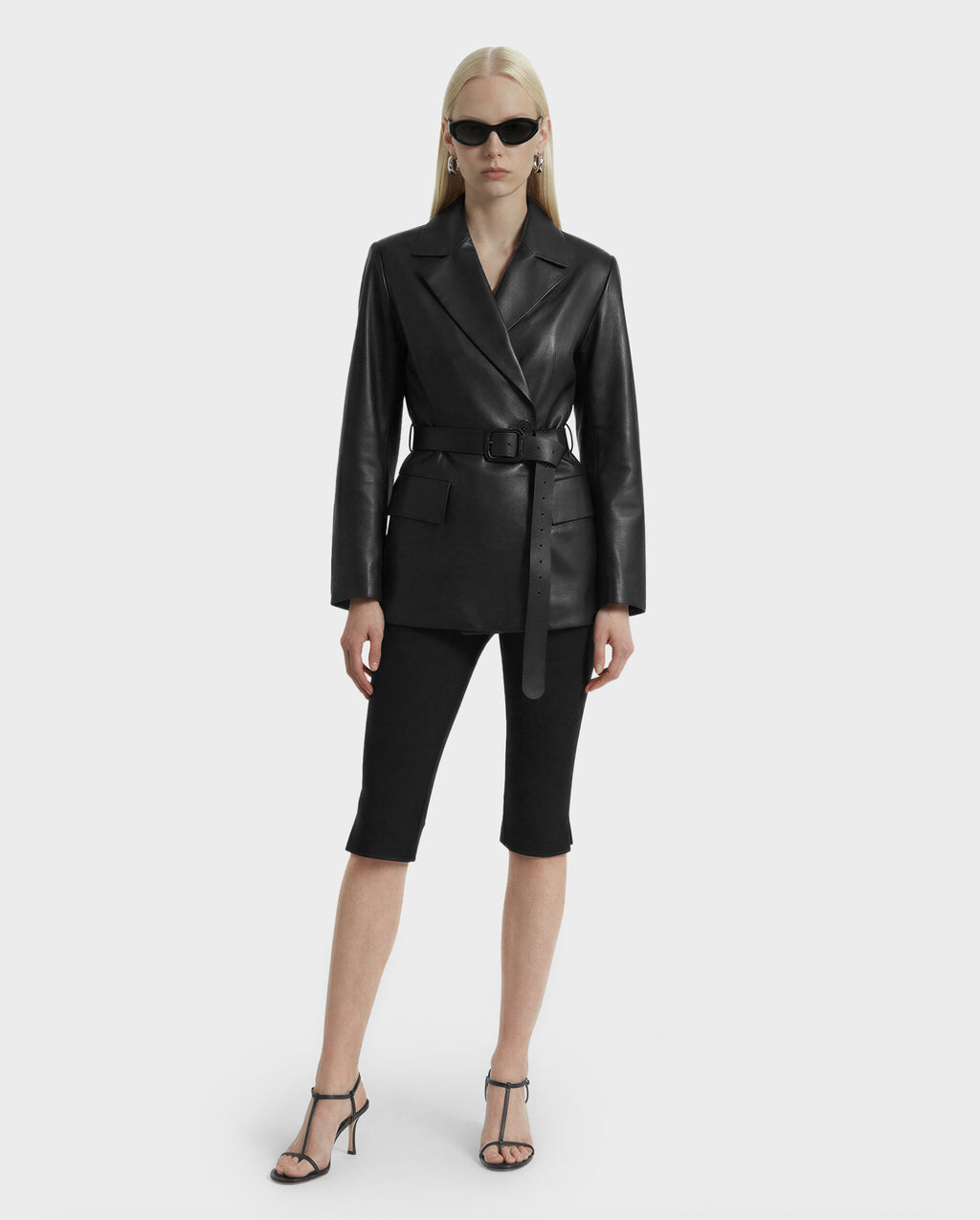A woman with long blonde hair wears Rudsak’s KENDALL - 8125617 Black leather blazer with a belt, black sunglasses, knee-length black shorts, and heeled sandals, standing against a plain white background.