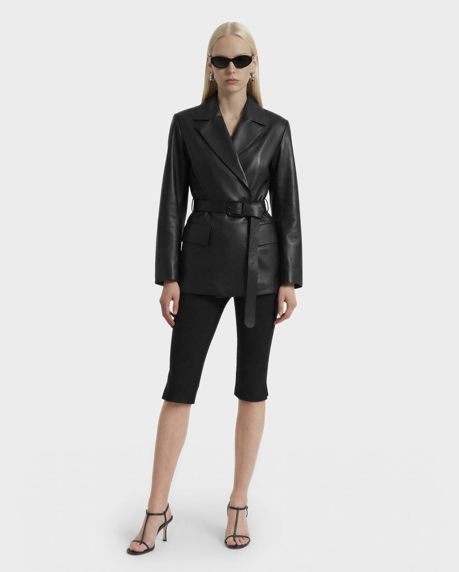 A woman with long blonde hair wears Rudsak’s KENDALL - 8125617 Black leather blazer with a belt, black sunglasses, knee-length black shorts, and heeled sandals, standing against a plain white background.
