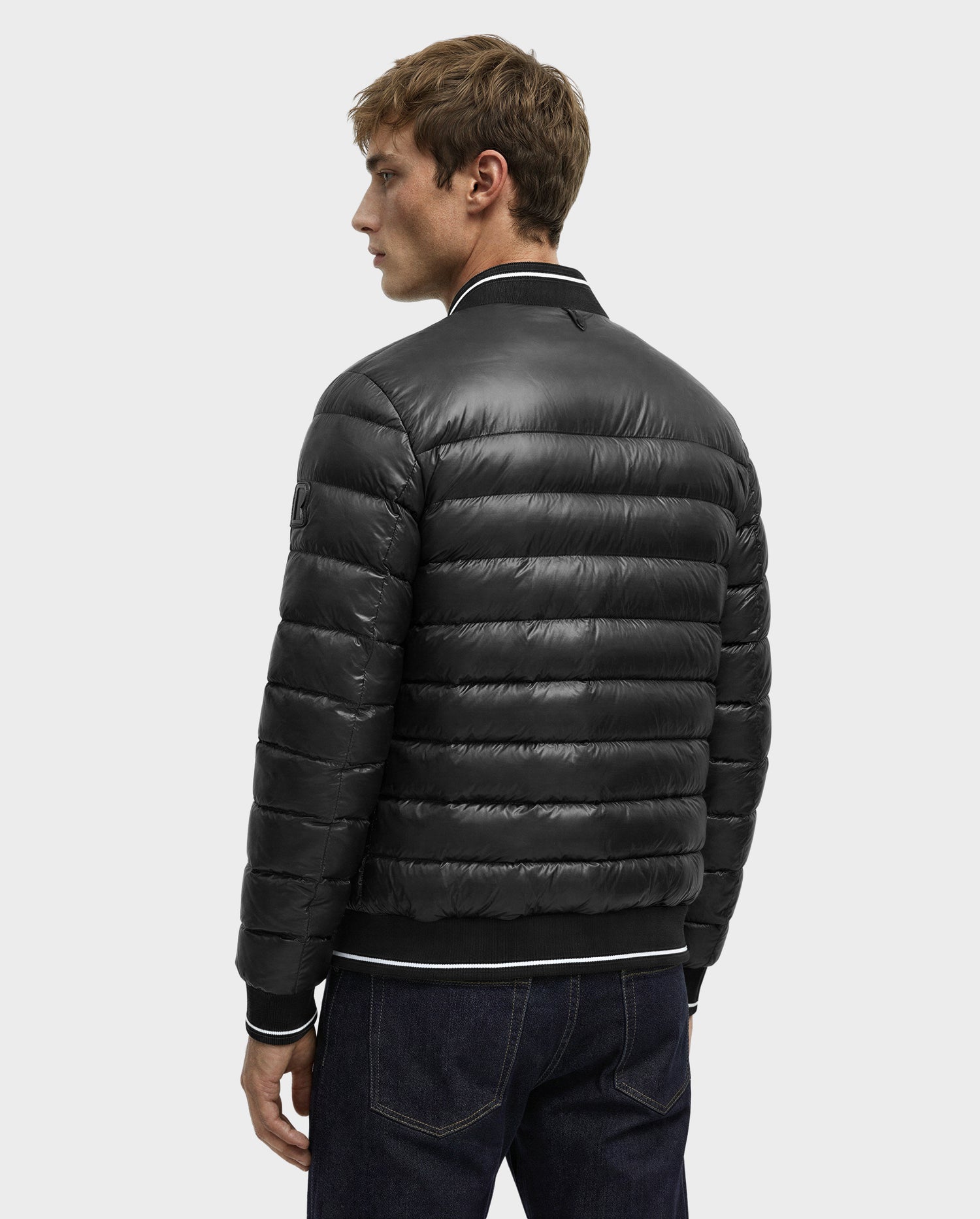 A person is shown from behind wearing the Rudsak JEREMY - 6125025 Black men’s down bomber jacket with quilted details and dark jeans against a plain light gray background.