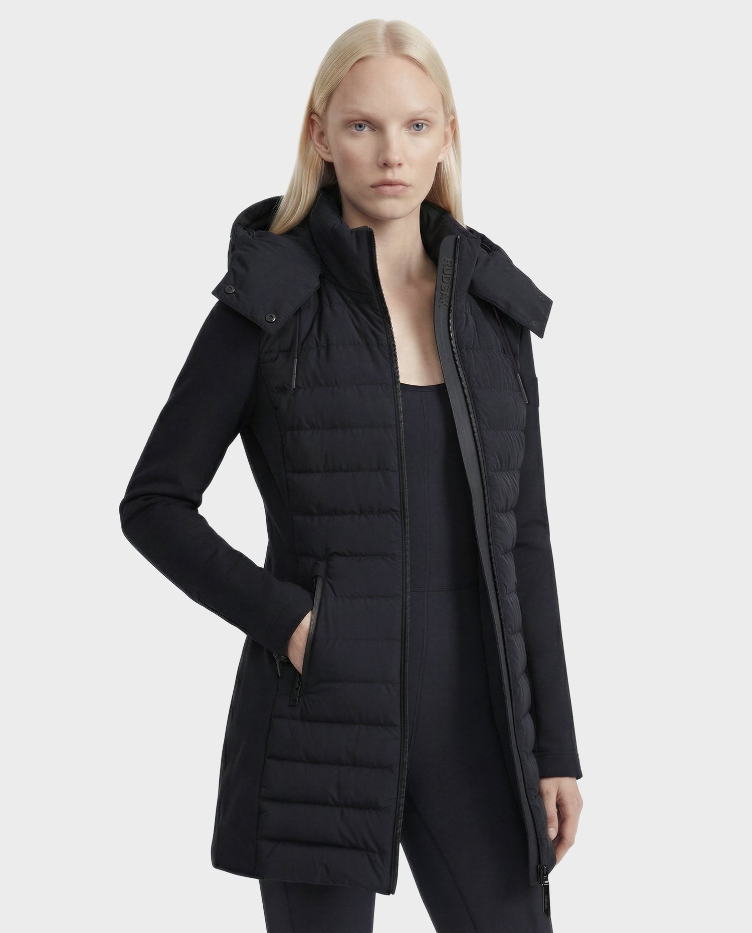 A woman with straight blonde hair models the Rudsak MERINA - 8125056 Black long quilted down jacket with a hood over a black top and pants, standing against a plain light gray background.