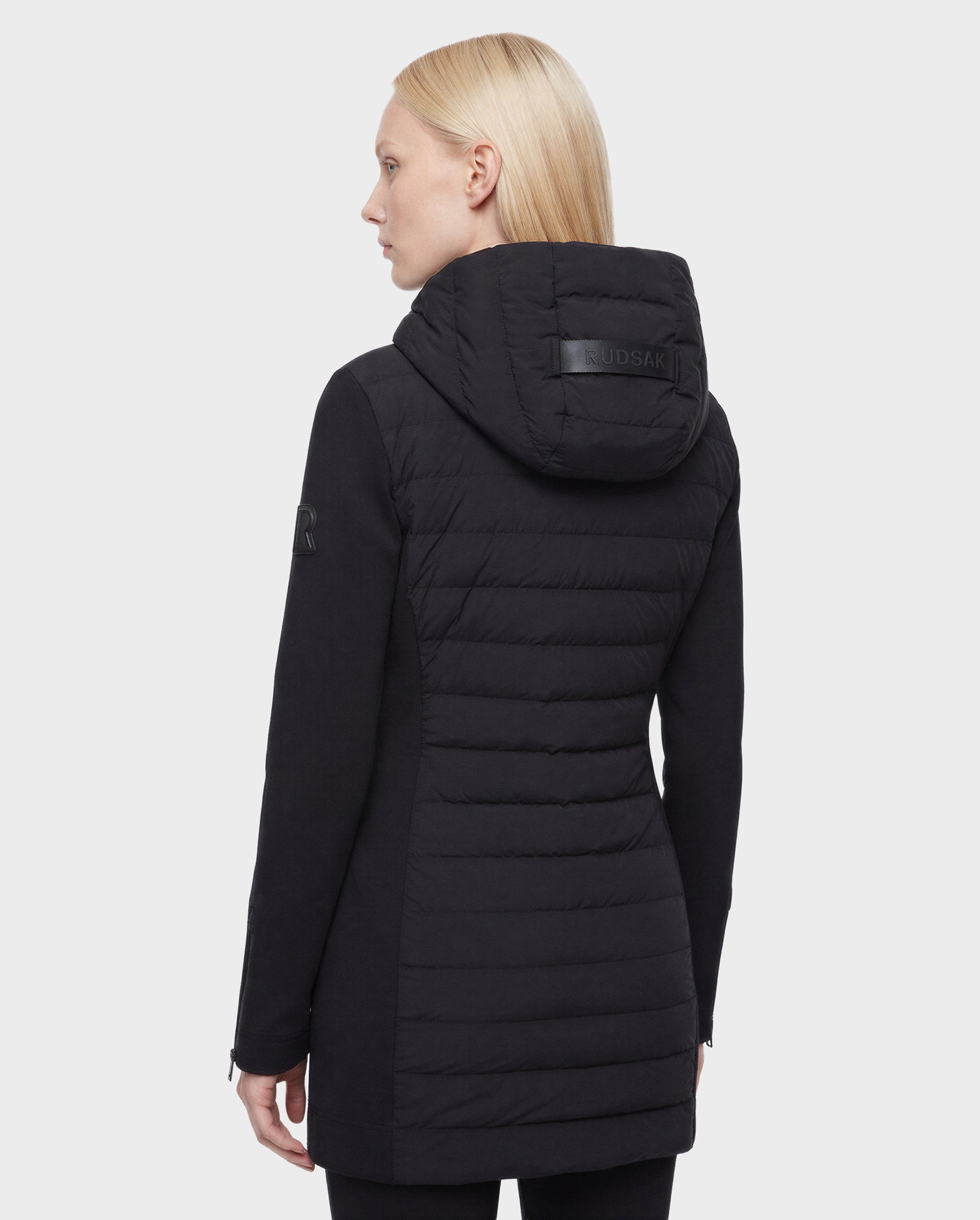 A person with straight blonde hair is shown from the back wearing the Rudsak MERINA - 8125056 Black quilted down jacket with RDS certified down, featuring logo patches on the hood and arm, against a plain light background.