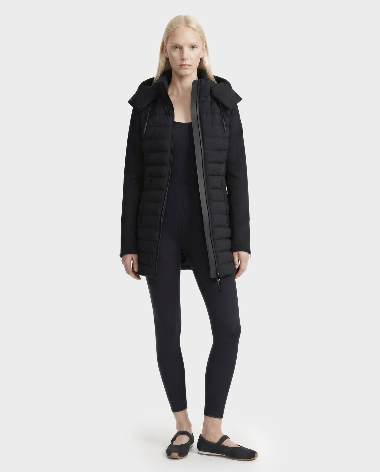A person with long blonde hair wears the Rudsak MERINA - 8125056 Black quilted down jacket over a black jumpsuit and black shoes, standing against a plain light gray background.