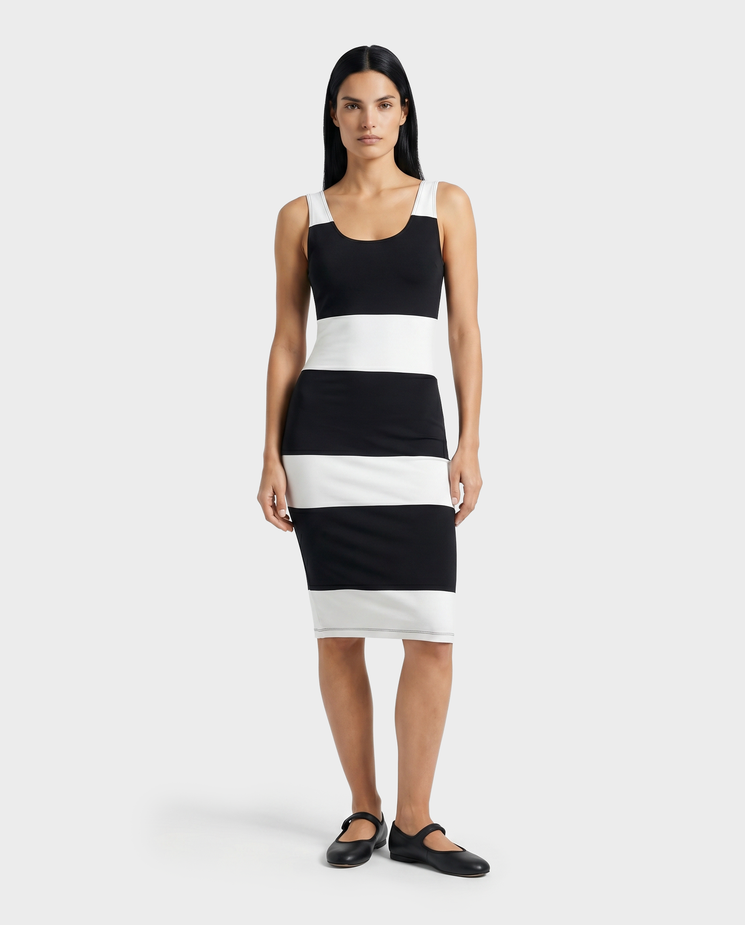 A woman faces forward in a sleeveless, knee-length DENISE - L523027 Optic White dress by Rudsak, featuring bold black and white horizontal stripes and black flats. The background is plain light gray.