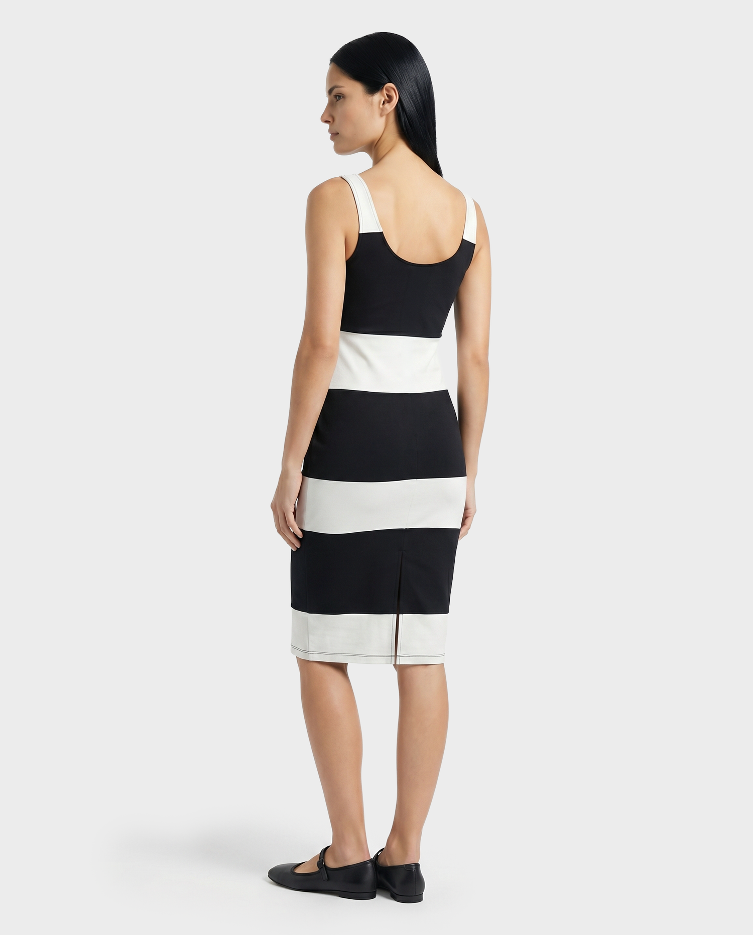 A woman with long dark hair faces away, wearing the Rudsak DENISE - L523027 Optic White sleeveless, knee-length dress with wide black and white stripes and black flats, set against a plain light background.