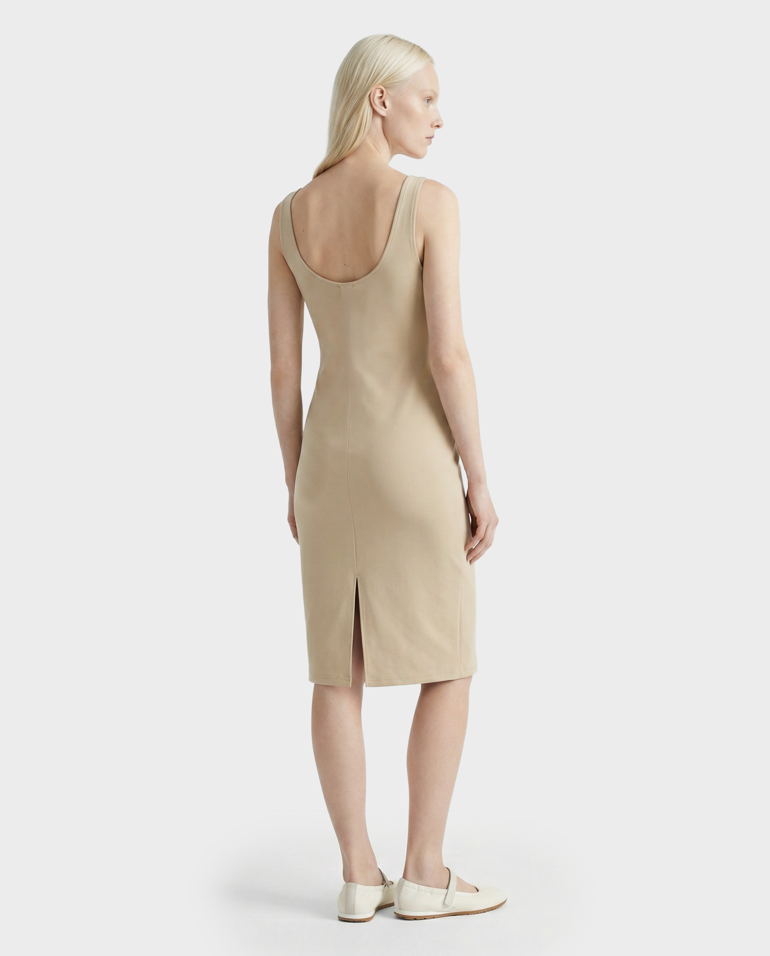 A woman with long, straight, blonde hair is wearing the Rudsak DENISE - L523027 Cream sleeveless, knee-length dress with a scoop back and small slit. She stands facing away from the camera in flat beige shoes.