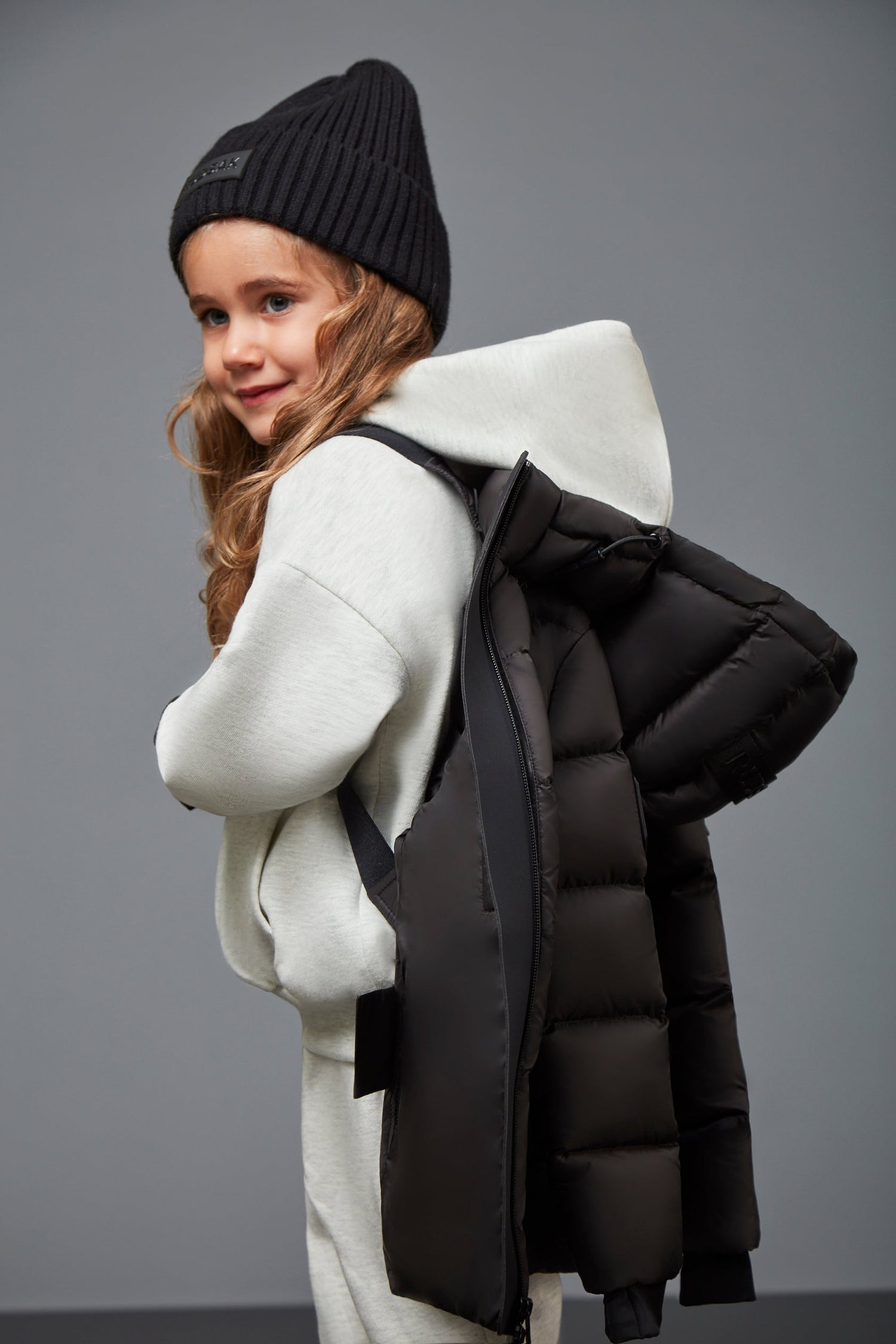 A young child smiles while carrying the Rudsak MILIA JR - BLACK K122503 kids down puffer jacket over their shoulder, pairing it with a gray hoodie and black knit beanie against a plain gray background—a true winter essential.