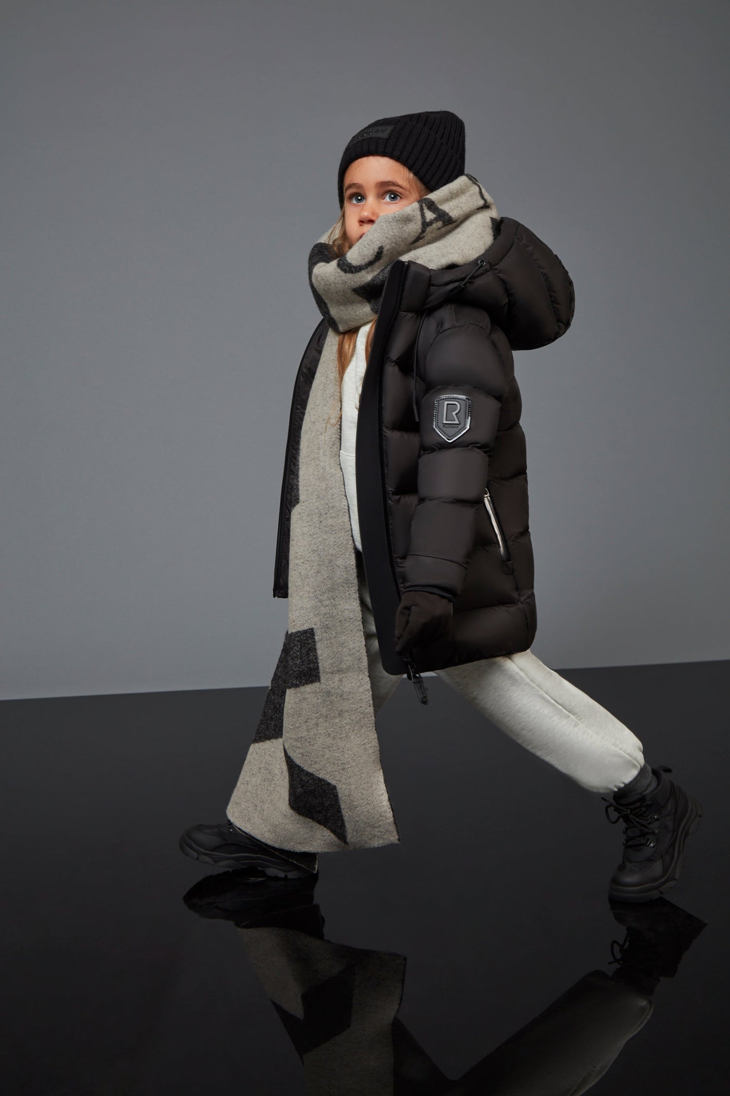 A child models the Rudsak MILIA JR - BLACK K122503 water-resistant jacket with a black hat, black boots, beige scarf, and white pants while walking confidently against a plain gray background.
