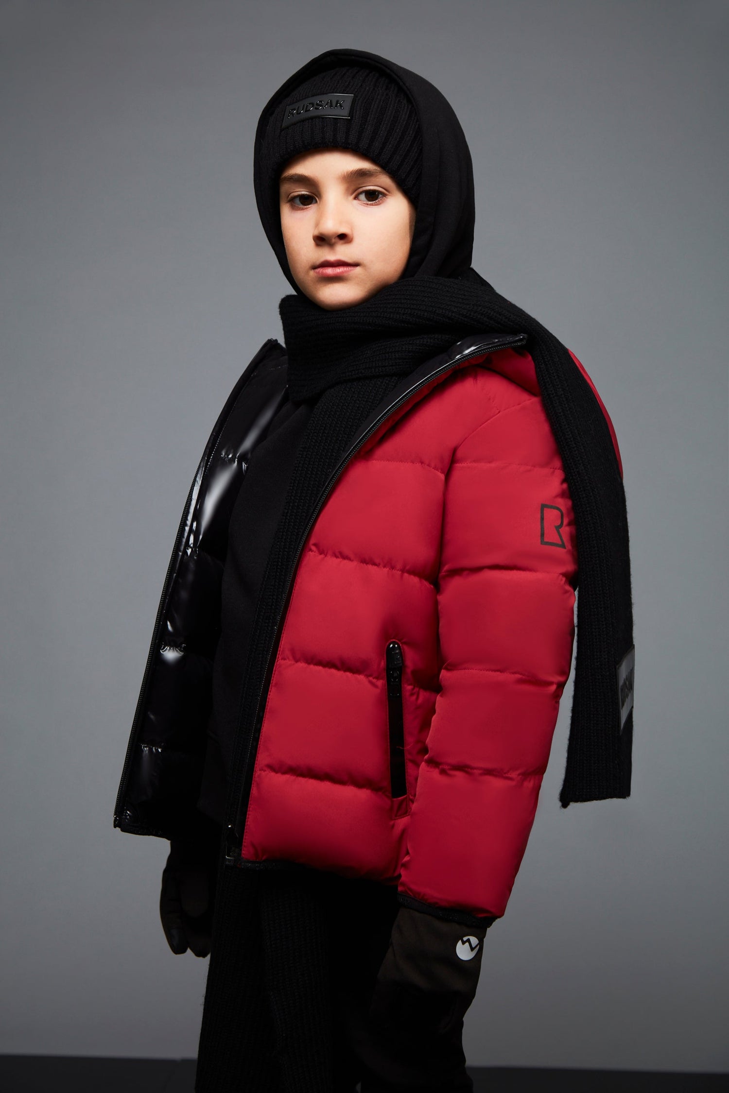 A young child confidently poses against a gray background wearing the Rudsak DEMIAN JR - K122500 red reversible down puffer, paired with a black scarf, gloves, hat, and dark pants.
