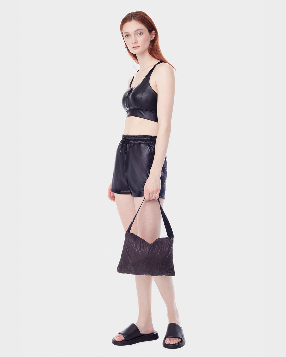 A woman with long red hair wears the RUDSAK NICKY - 9123005 BLACK F vegan leather shoulder bag, paired with a black crop top, black shorts, and black slide sandals against a plain white background.