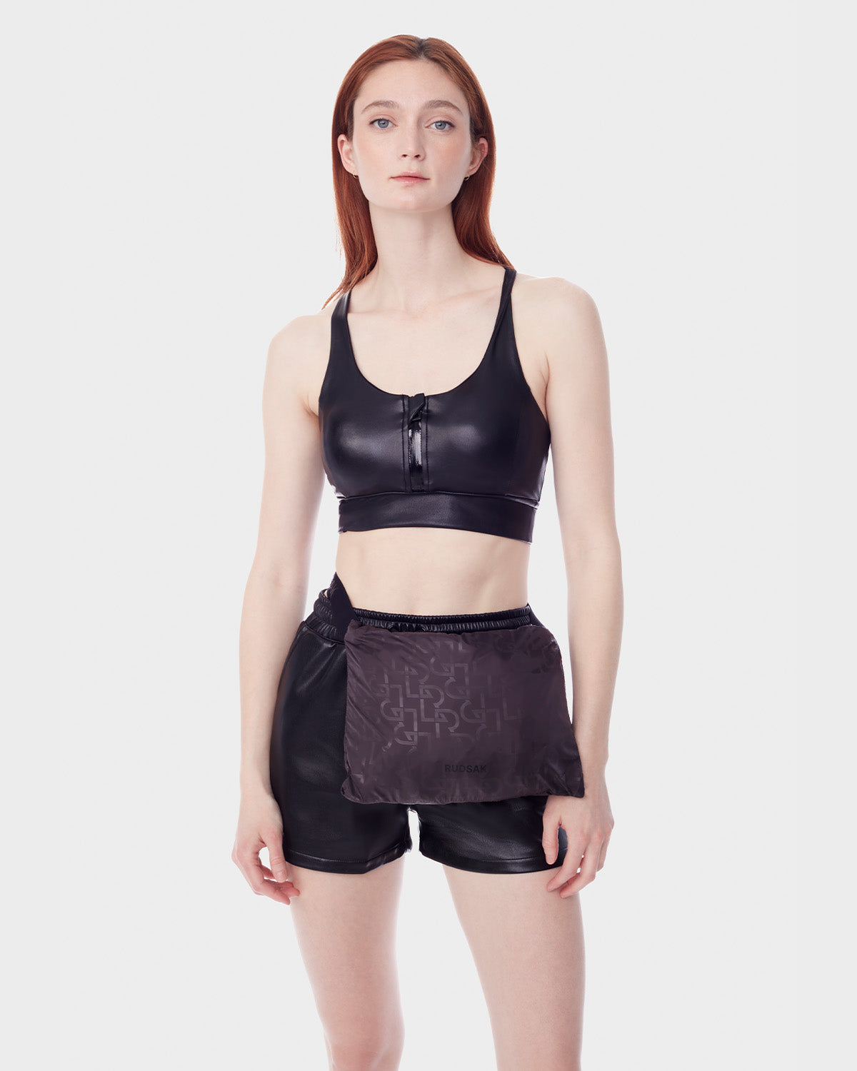 A woman with straight red hair wears the RUDSAK NICKY - 9123005 BLACK F vegan leather-look crop top, paired with black shorts and a black belt bag, standing against a plain white background.
