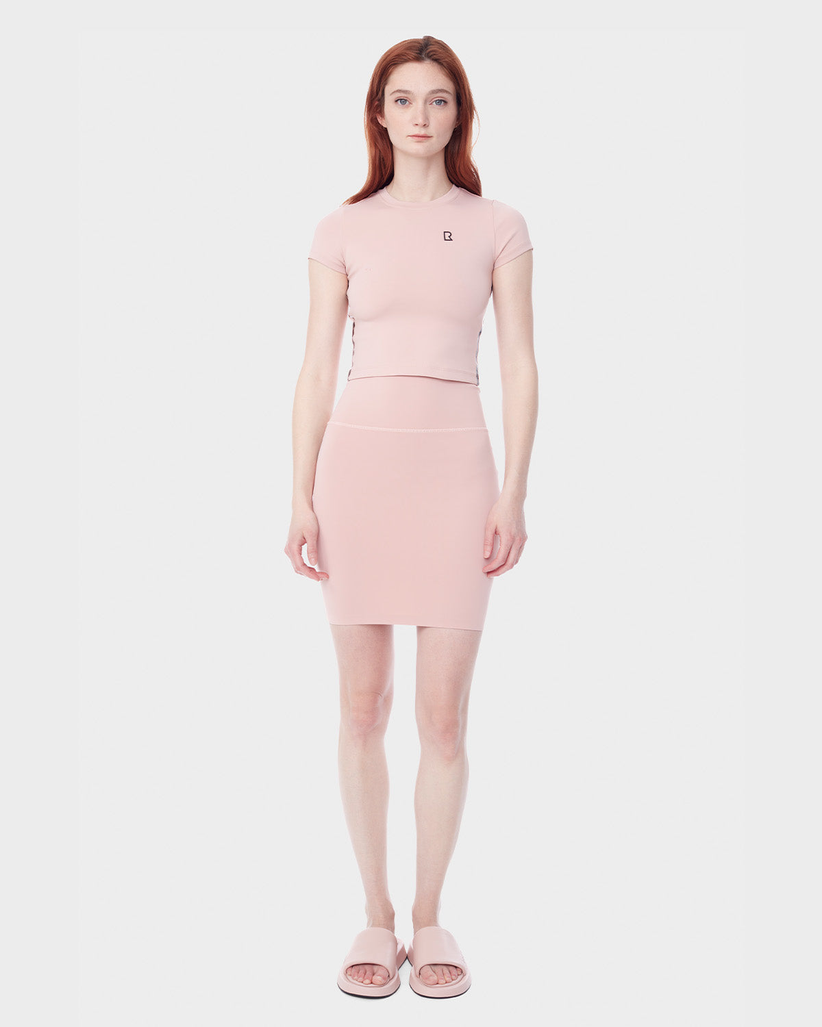 A woman with long red hair models the Rudsak JESS - 8523042 BLUSH fitted light pink top and matching high-waisted skirt, paired with pink slide sandals, standing against a plain white background—ideal for refreshing your spring wardrobe.