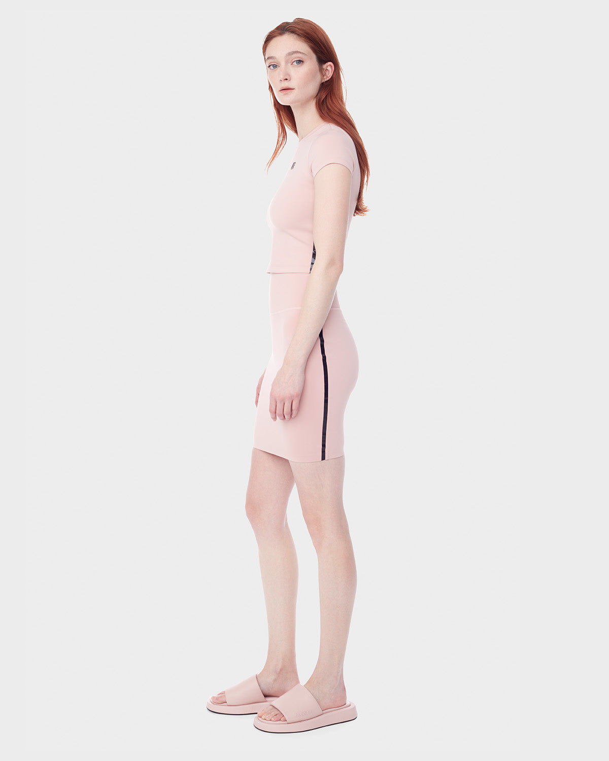A woman with long red hair stands sideways wearing the Rudsak JESS - 8523042 BLUSH fitted light pink crop top and matching mini skirt with black side stripes, paired with light pink slide sandals—the perfect spring look.