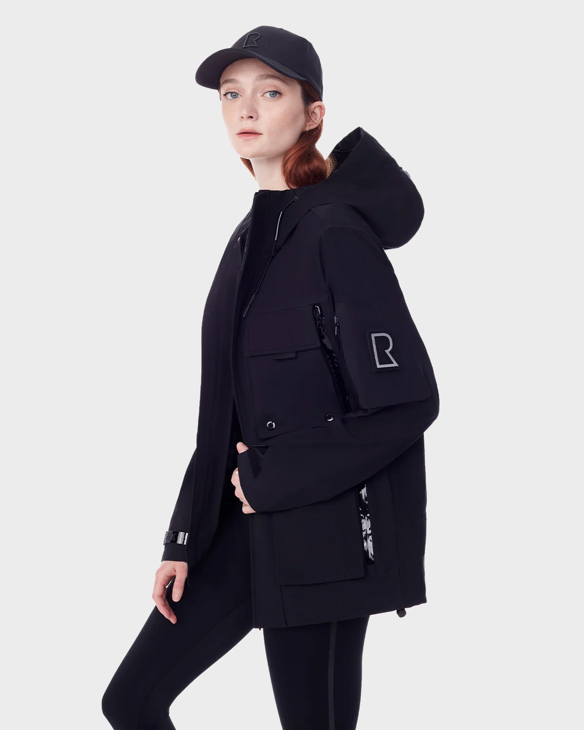 A woman wears the RUDSAK PALM - 9123000 BLACK F unisex rainwear jacket made from recycled stormshell fabric, featuring a large R sleeve patch and multiple pockets, with a black cap and leggings against a light gray background.