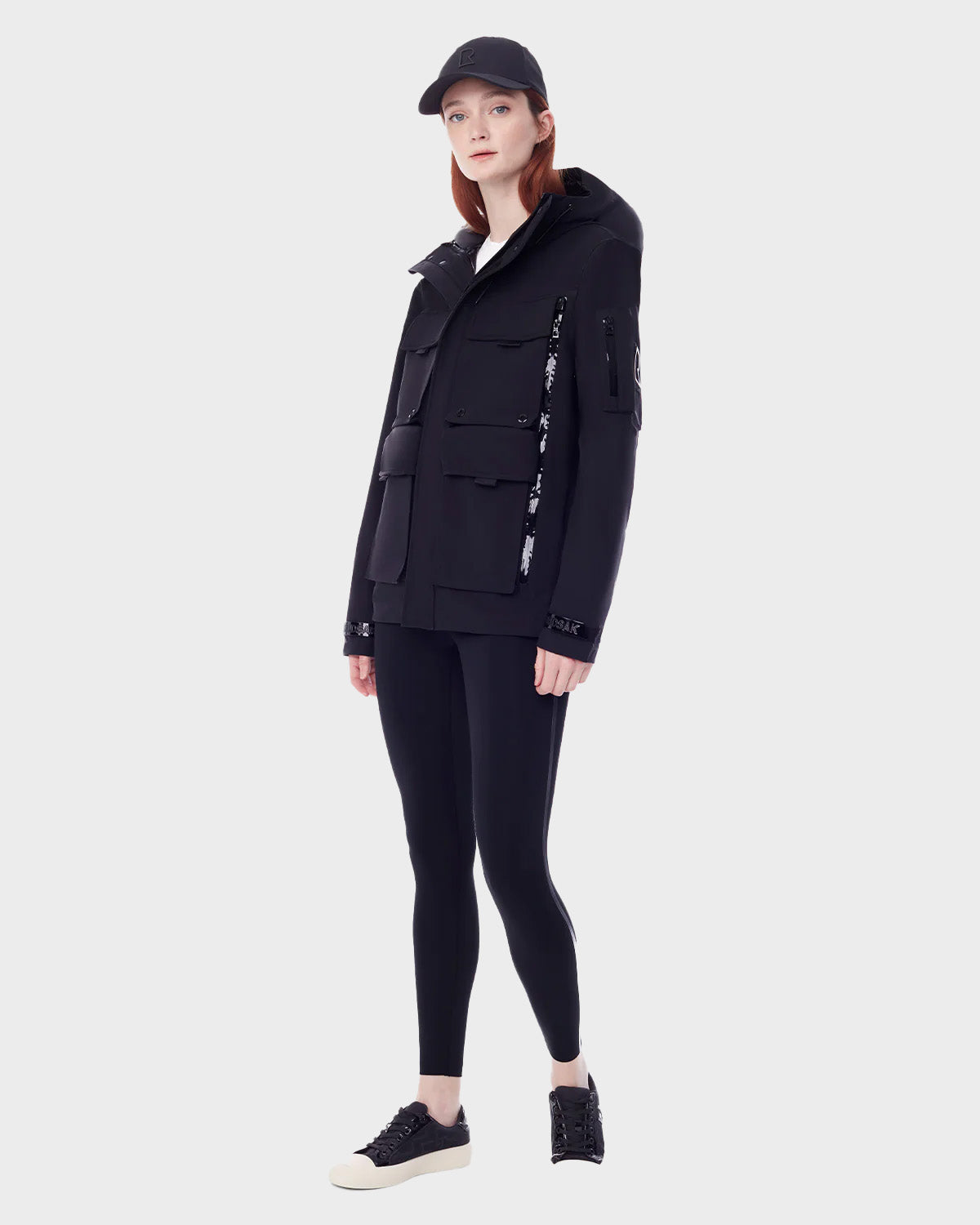 A woman in the RUDSAK PALM - 9123000 BLACK F utility rainwear jacket, paired with black leggings and black sneakers with white soles, stands against a plain light gray background.