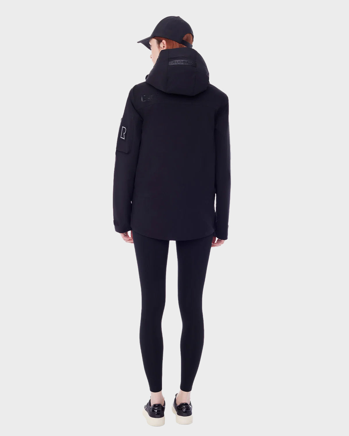 A person faces away, wearing the RUDSAK PALM - 9123000 BLACK F unisex utility rainwear jacket, black leggings, a black cap, and black sneakers with white soles, set against a plain light gray background.