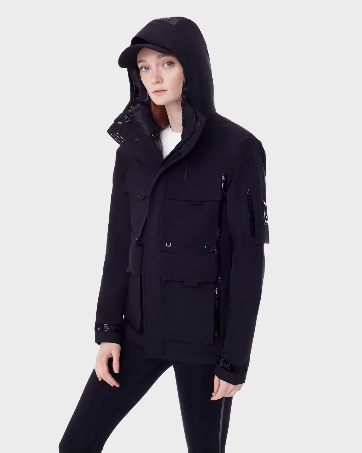 A woman in the RUDSAK PALM - 9123000 BLACK F utility rainwear jacket, black cap, and black pants stands against a light gray background, gazing slightly to the side with a neutral expression.