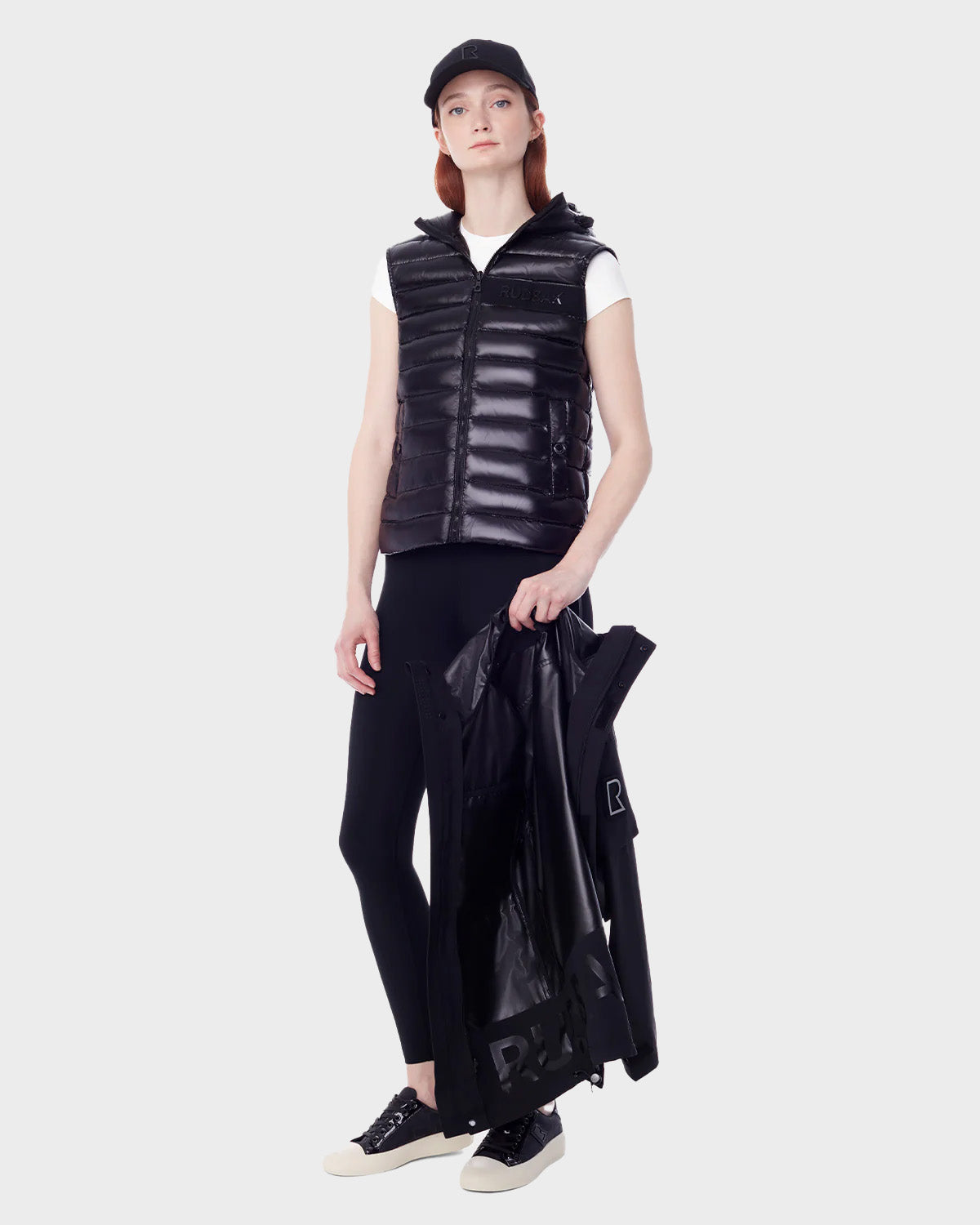 A woman stands against a plain background wearing the RUDSAK PALM - 9123000 BLACK F cap, black puffer vest over a white t-shirt, black leggings, and black-and-white sneakers, holding an RLX unisex utility rainwear jacket in her left hand.
