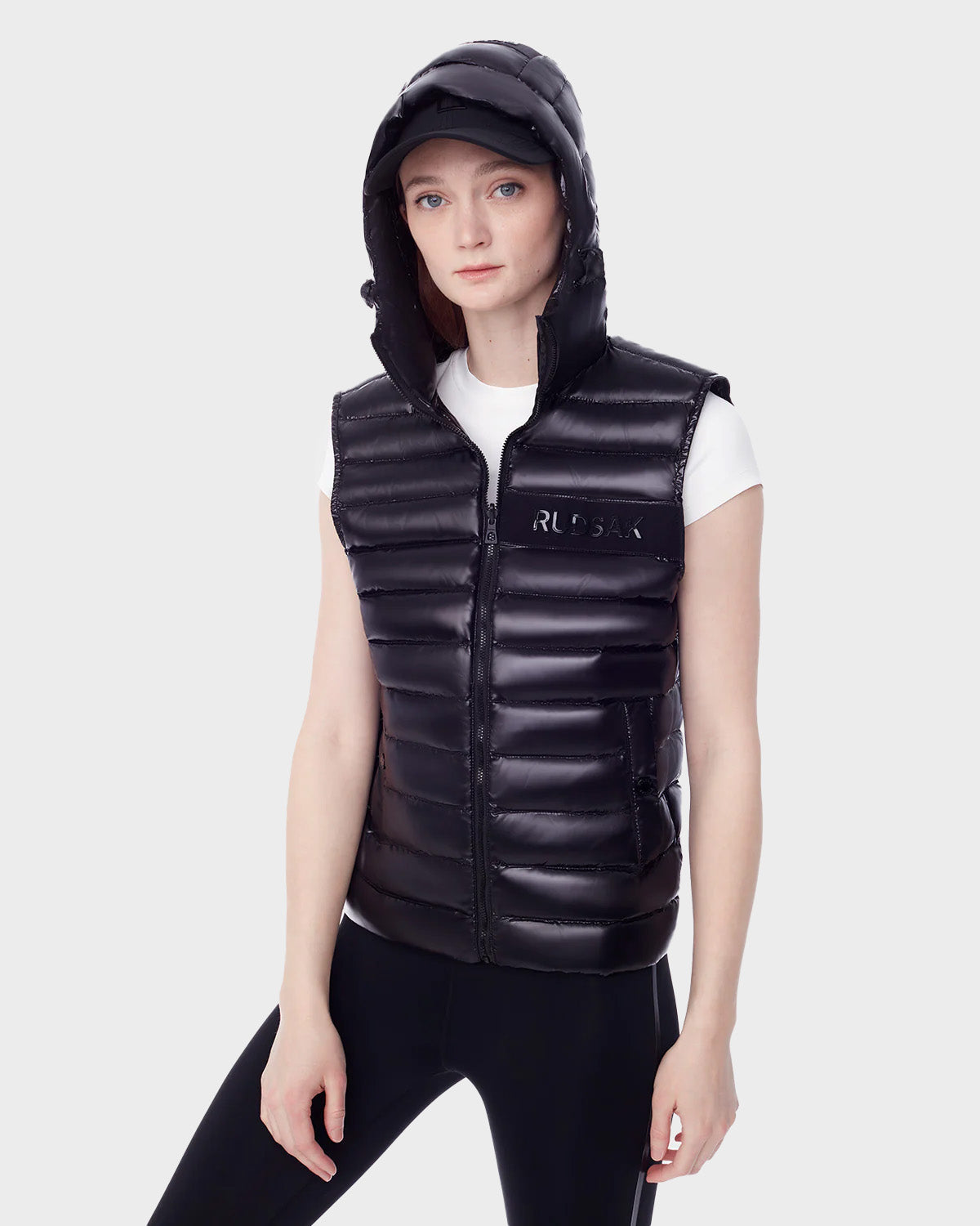 A woman wears the RUDSAK PALM - 9123000 BLACK F shiny black hooded puffer vest, crafted from recycled stormshell fabric, paired with a black cap, white short-sleeve shirt, and black leggings against a plain white background.