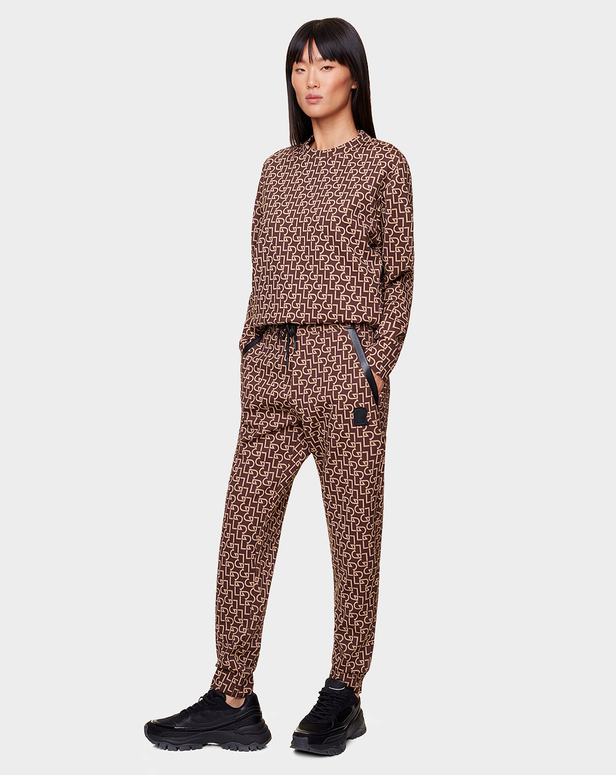 A woman with long dark hair stands against a light gray background, wearing the OLAV - 9522503 RUDSAK PATTERN WOMEN relaxed-fit geometric tracksuit with recycled leather trims by Rudsak, hands in pockets.