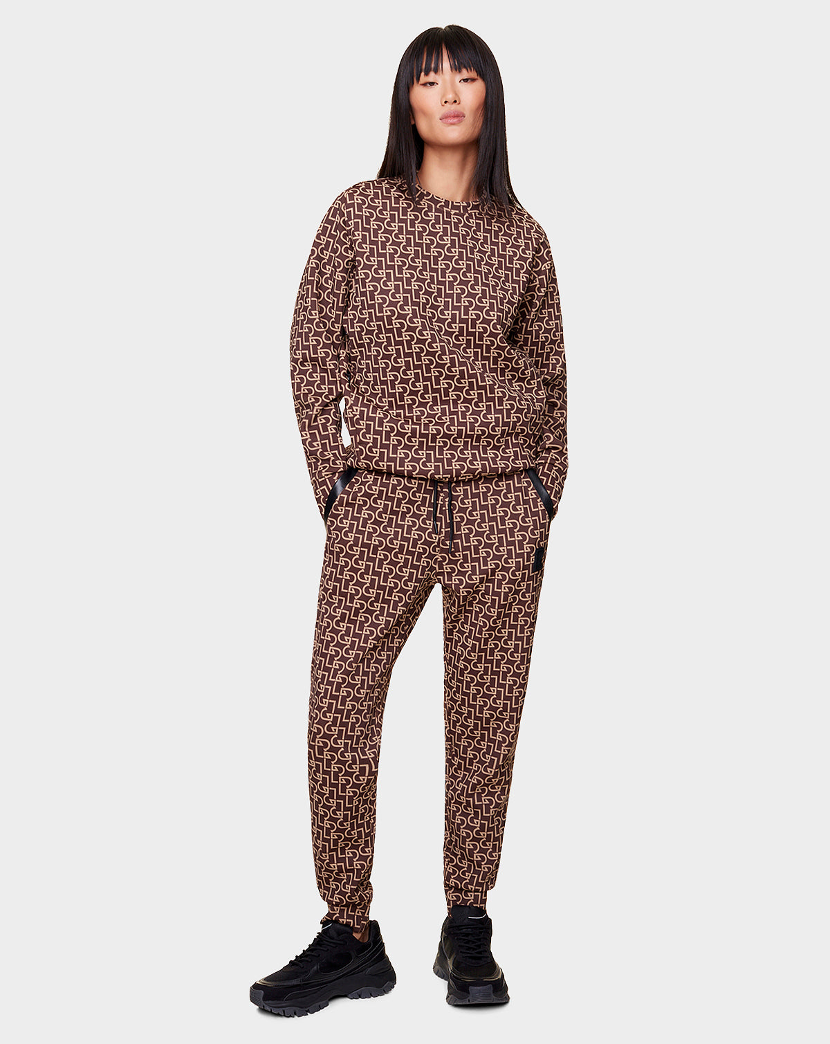 A woman stands with hands in pockets, wearing Rudsaks OLAV - 9522503 RUDSAK PATTERN WOMEN joggers and matching sweatshirt with a beige geometric pattern and recycled leather trims, paired with black sneakers, against a plain light background.