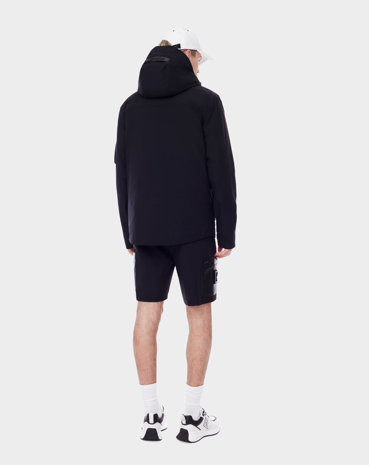 A person faces away wearing RUDSAK's PALM - 9123000 BLACK unisex rainwear jacket made from recycled Stormshell fabric, styled with black shorts, white socks and sneakers, and a white cap against a plain light background.