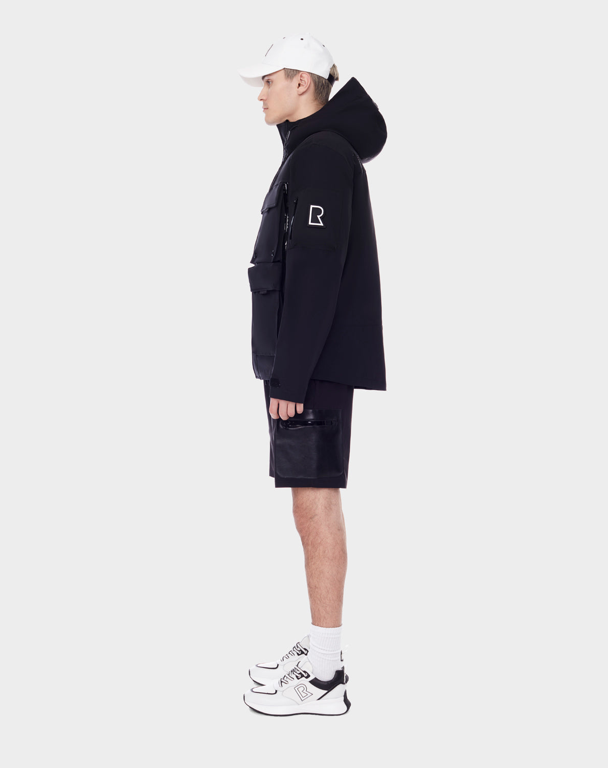 A person stands in profile against a plain white background, wearing the RUDSAK PALM - 9123000 BLACK unisex utility rainwear jacket, black shorts with a pocket, white socks, white sneakers, and a white baseball cap.