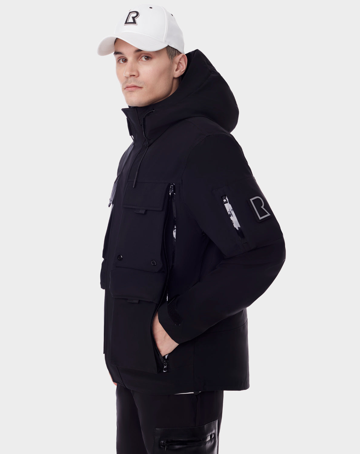 A man stands sideways in the PALM - 9123000 BLACK utility rainwear jacket by RUDSAK, featuring multiple pockets, a bold R logo, and unisex fit. He wears a white cap with the same logo. The recycled Stormshell jacket is shown on a plain white background.