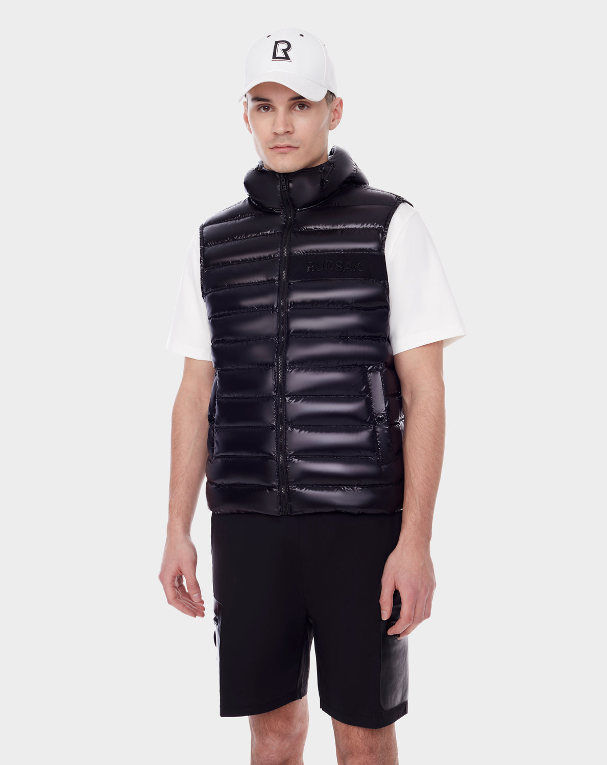 A man stands against a plain white background wearing RUDSAK's PALM - 9123000 BLACK: a shiny black puffer vest with hood in recycled Stormshell fabric, paired with a white baseball cap, short-sleeve shirt, and black shorts.