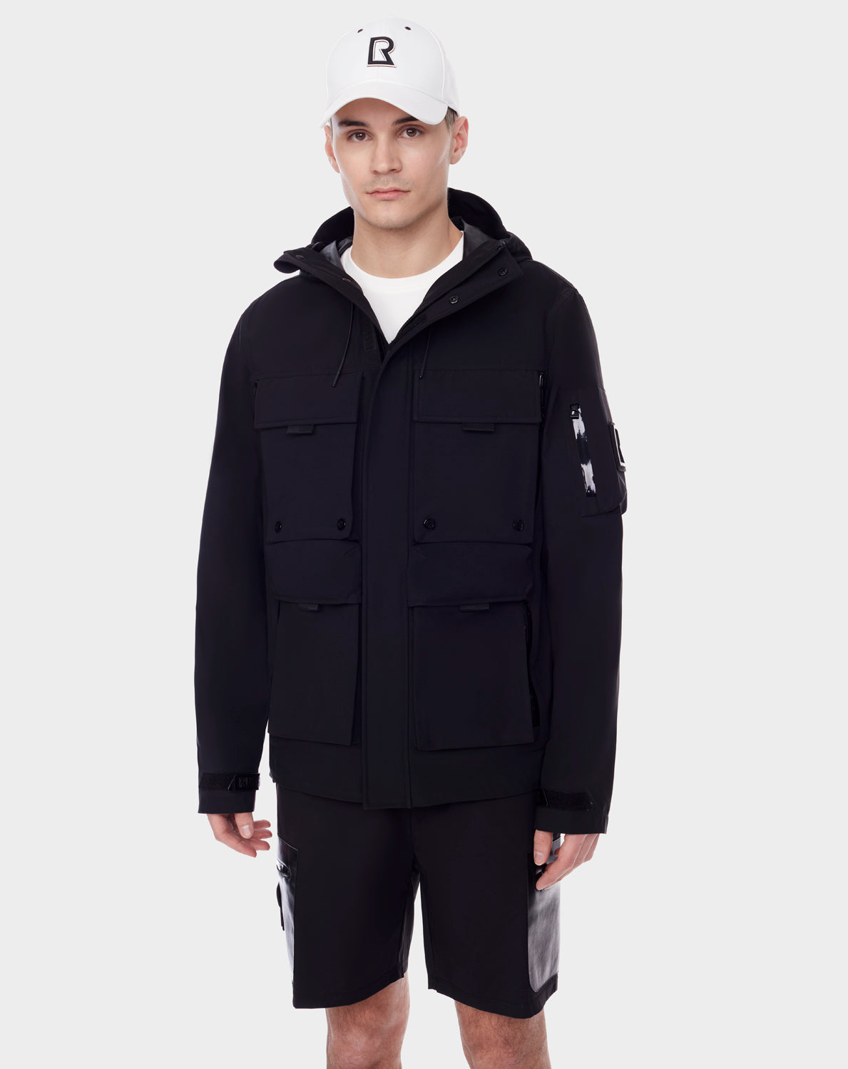 A young man wears the RUDSAK PALM - 9123000 BLACK unisex utility rainwear jacket with multiple front pockets, paired with a white baseball cap and black shorts, standing against a plain white background.