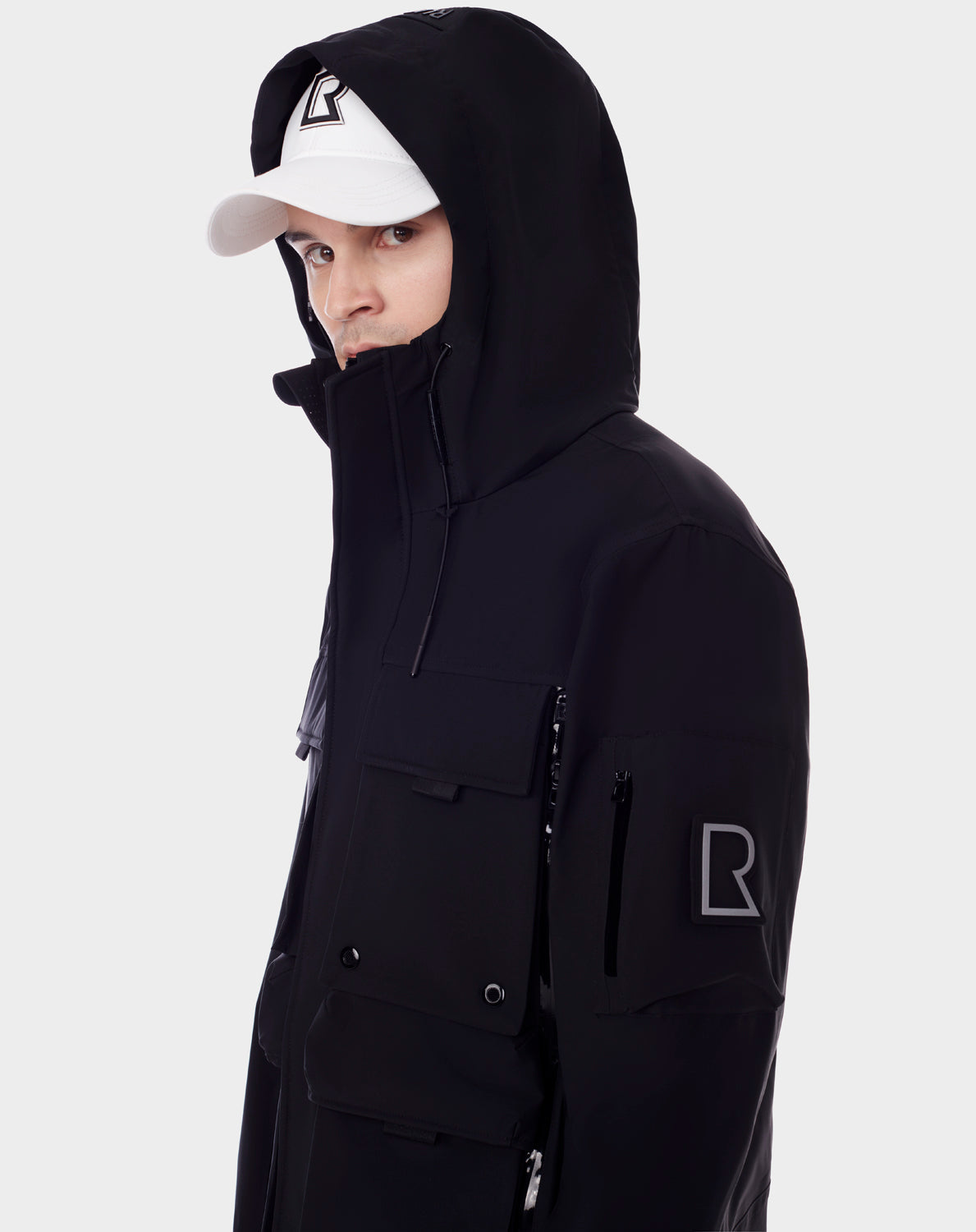 Wearing RUDSAK’s PALM - 9123000 BLACK rain jacket with multiple pockets and an R logo patch, plus a white cap featuring a bold R logo, a person faces the camera against a plain light gray background. The jacket features recycled Stormshell fabric.