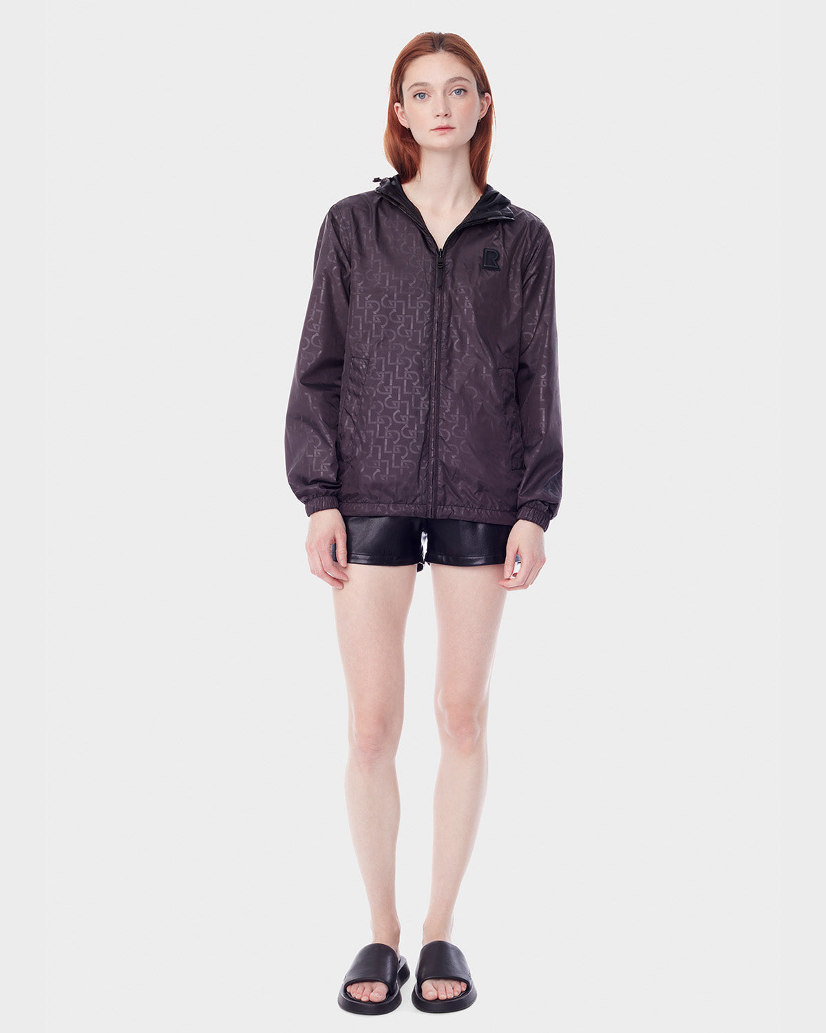 A woman with straight, shoulder-length hair faces forward wearing RUDSAK's NICKY - 9123005 BLACK F reversible lightweight jacket, black shorts, and black slide sandals against a plain white background.