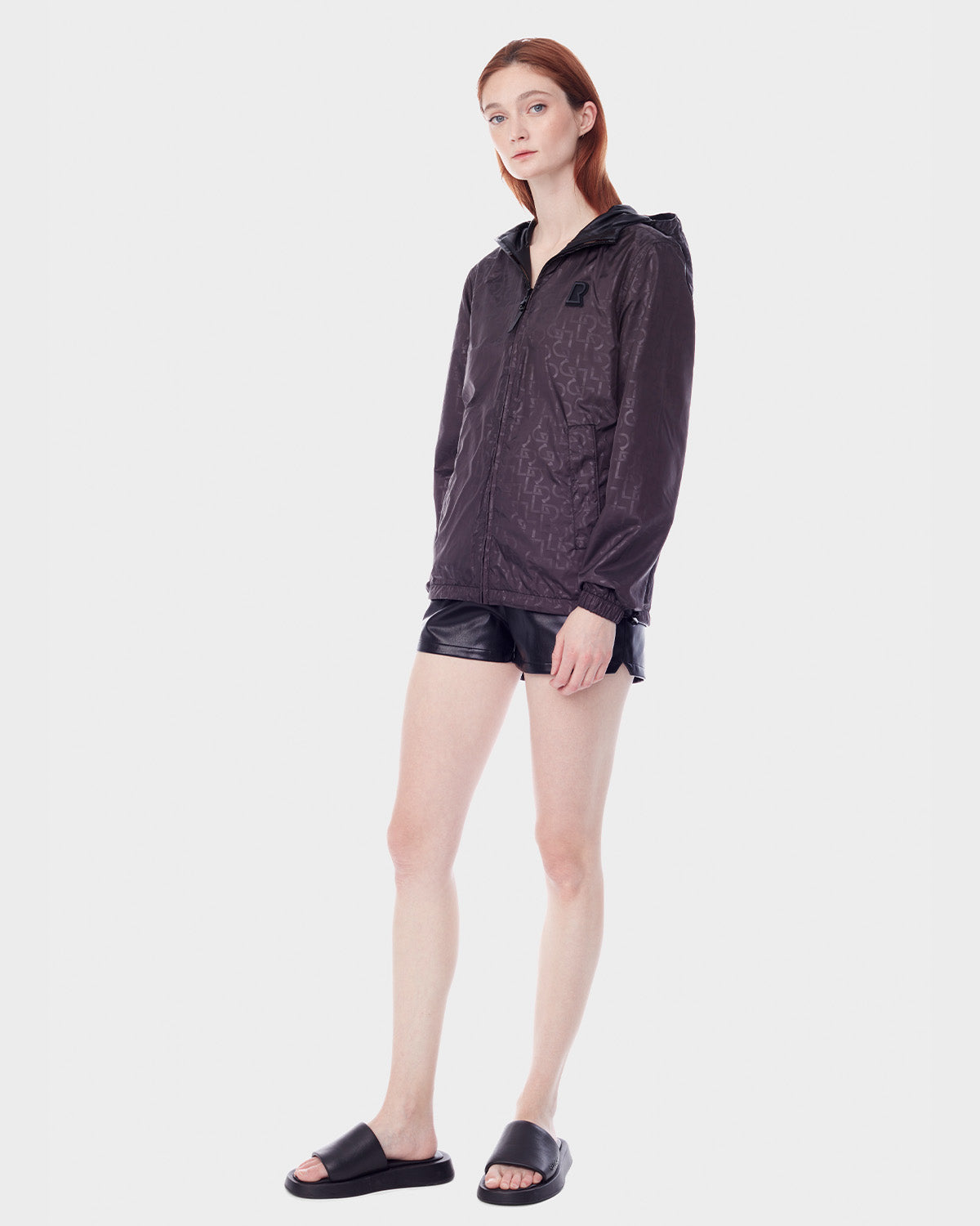 A woman with straight, reddish-brown hair and a neutral expression models the RUDSAK NICKY - 9123005 BLACK F unisex windbreaker against a plain white background, paired with black shorts and slide sandals.
