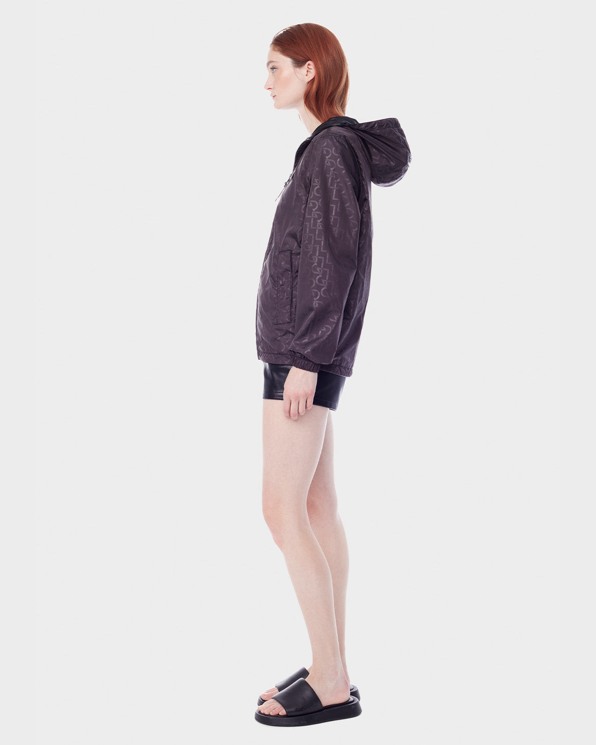 A person with long red hair stands in profile, wearing the RUDSAK NICKY - 9123005 BLACK F unisex windbreaker, black shorts, and black slide sandals against a plain white background.