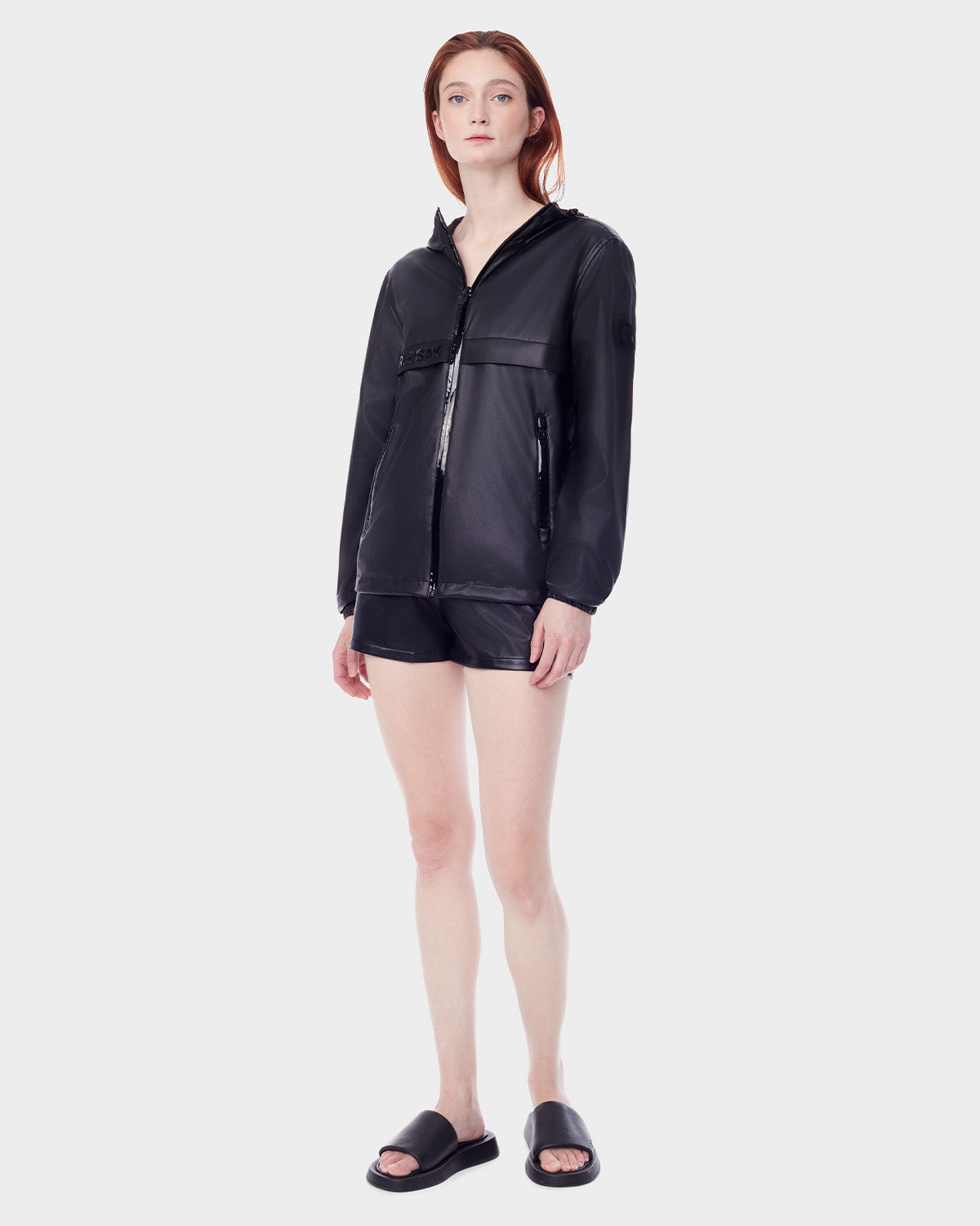 A woman with straight, shoulder-length hair wears the RUDSAK NICKY - 9123005 BLACK F reversible lightweight jacket, black shorts, and black slide sandals. She stands slightly angled against a plain white background.