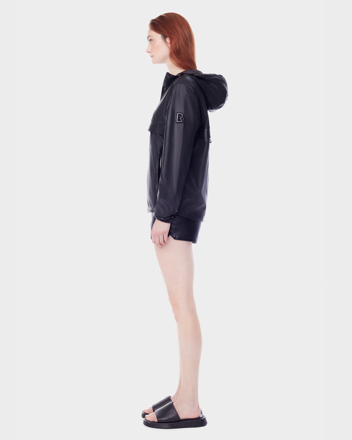 A person with long red hair stands in profile wearing the RUDSAK NICKY - 9123005 BLACK F unisex windbreaker, black shorts, and black slide sandals against a plain white background.