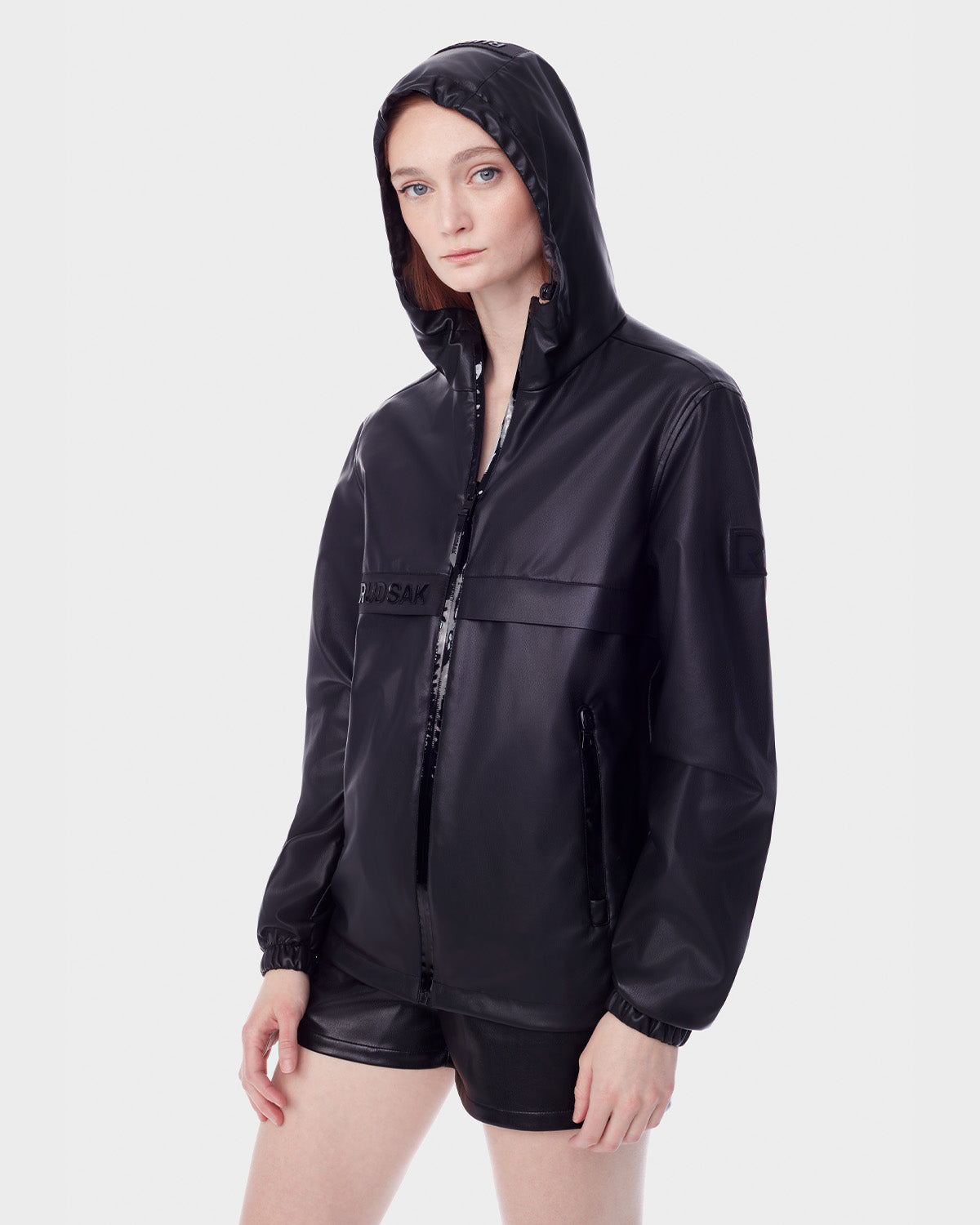 A person models the RUDSAK NICKY - 9123005 BLACK F unisex vegan leather windbreaker with black shorts, standing against a plain white background and facing the camera with a neutral expression.