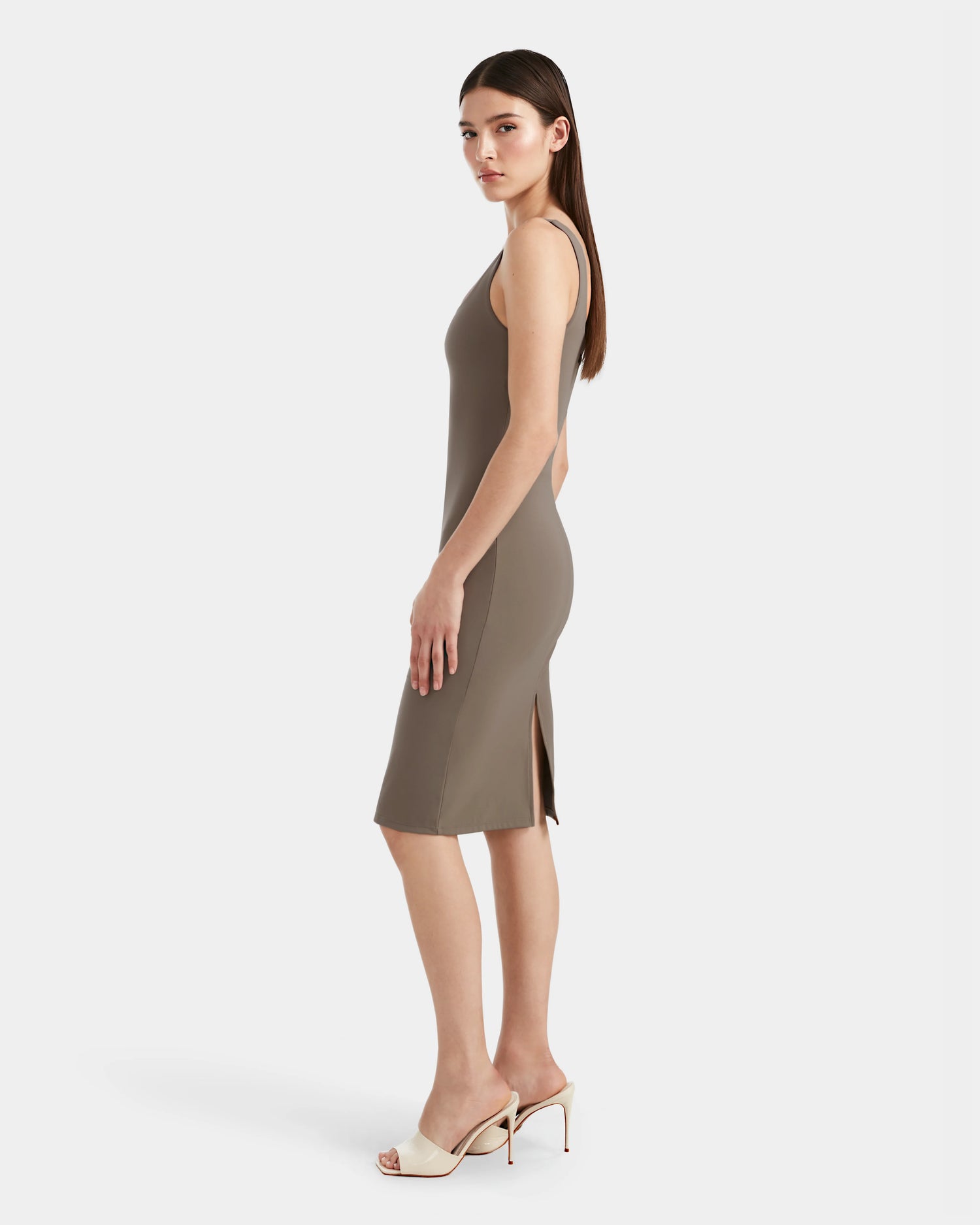 A woman stands in profile wearing the Rudsak SIMONE - 8524065 Cement women's midi dress with a side slit, crafted from stretch performance fabric, paired with white heeled sandals, set against a plain white background.