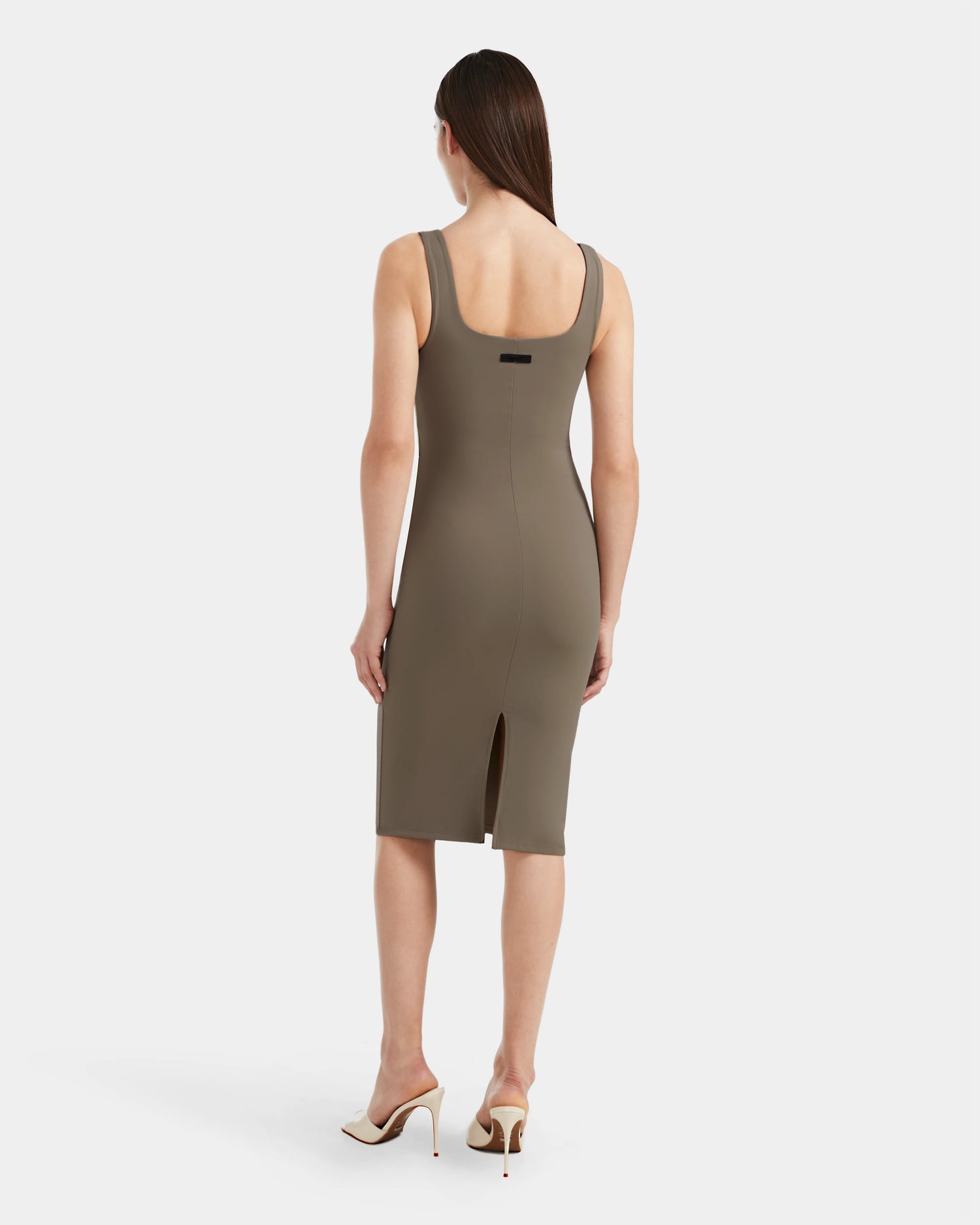 A woman with straight brown hair wears the Rudsak SIMONE - 8524065 Cement fitted sleeveless midi dress in taupe, knee-length with a back slit, paired with white heels, standing against a plain white background.