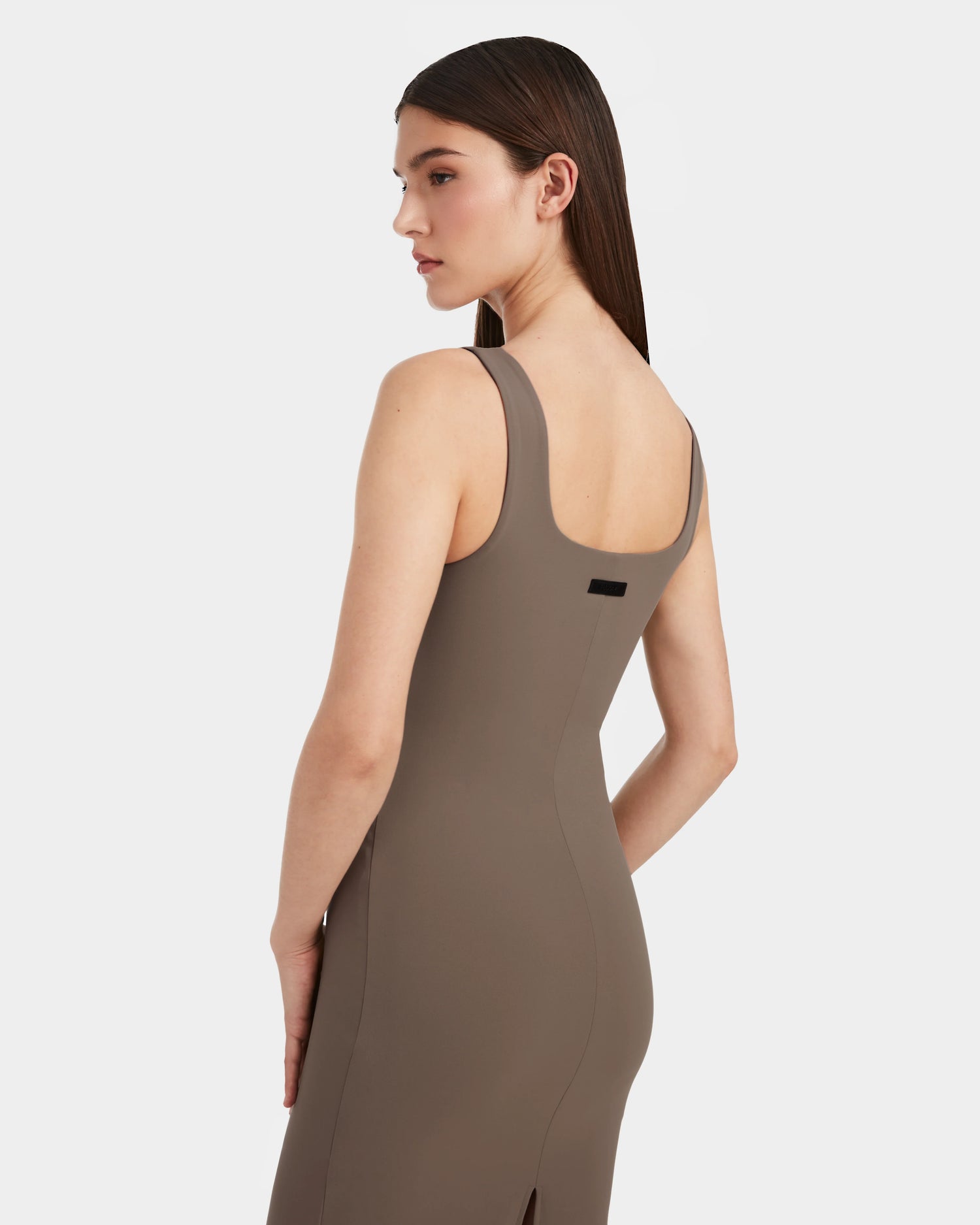 A woman with straight brown hair models the Rudsak SIMONE - 8524065 Cement sleeveless taupe midi dress, standing slightly turned and looking over her shoulder against a white background.