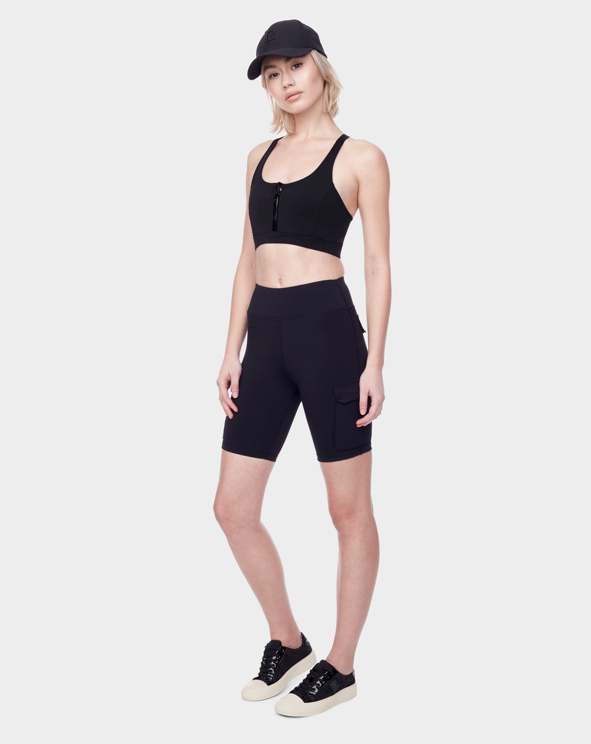 A woman stands against a white background wearing the Rudsak SERINA - 8521006 BLACK sneakers with white soles, paired with a black sports bra, black high-waisted biker shorts with a side pocket, and a black baseball cap.