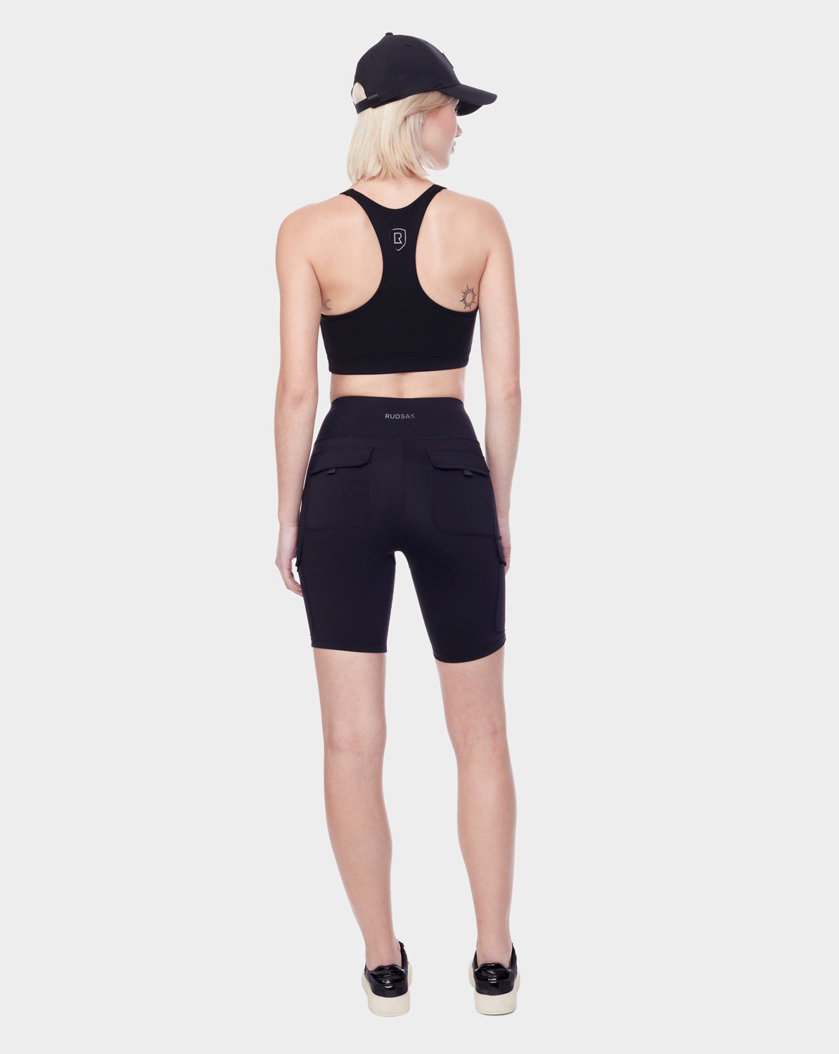 A woman with short blonde hair and a small tattoo on her left arm wears Rudsak's SERINA - 8521006 BLACK biker shorts, featuring cargo-style pockets and high-performance stretch fabric, paired with a matching sports bra, sneakers, and a cap.