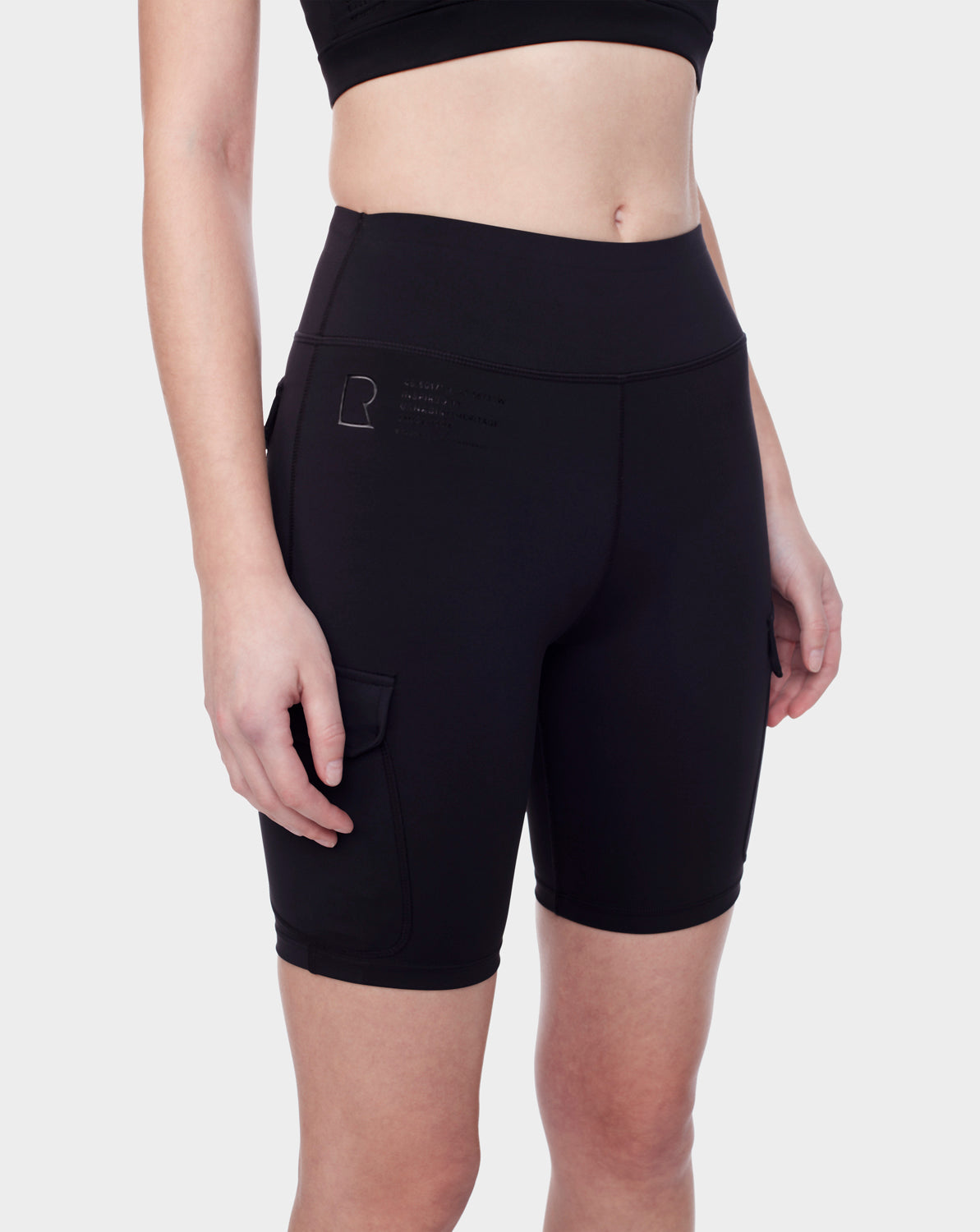 A person models Rudsak’s SERINA - 8521006 BLACK high-waisted biker shorts for women, featuring a side pocket and small logo on the left hip. Photo is cropped shoulders to knees against a plain light background.